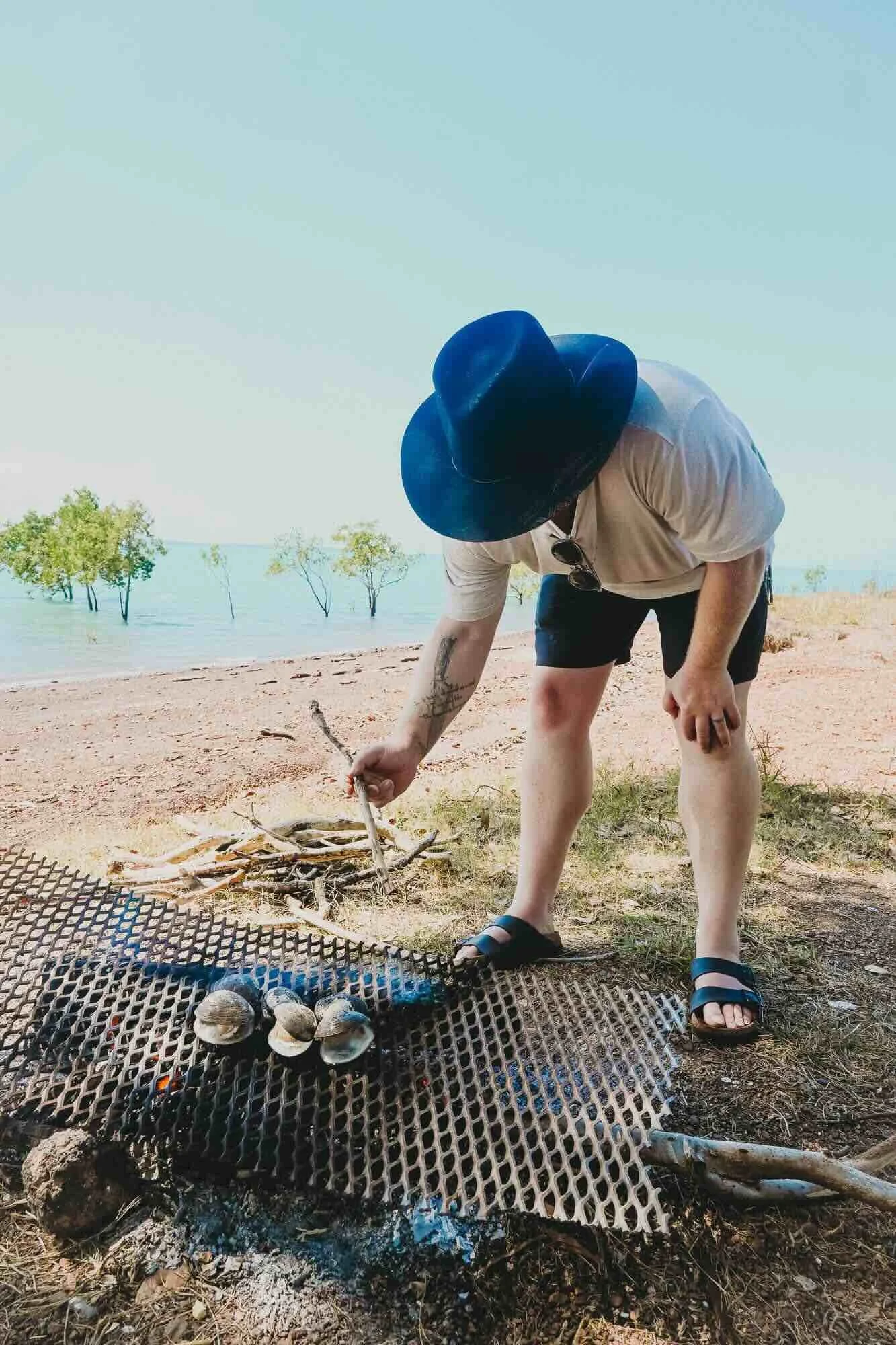 A person wearing a large black hat, white T-shirt, black shorts, and sandals, is tending to a small campfire on a beach, surrounded by lake and trees.