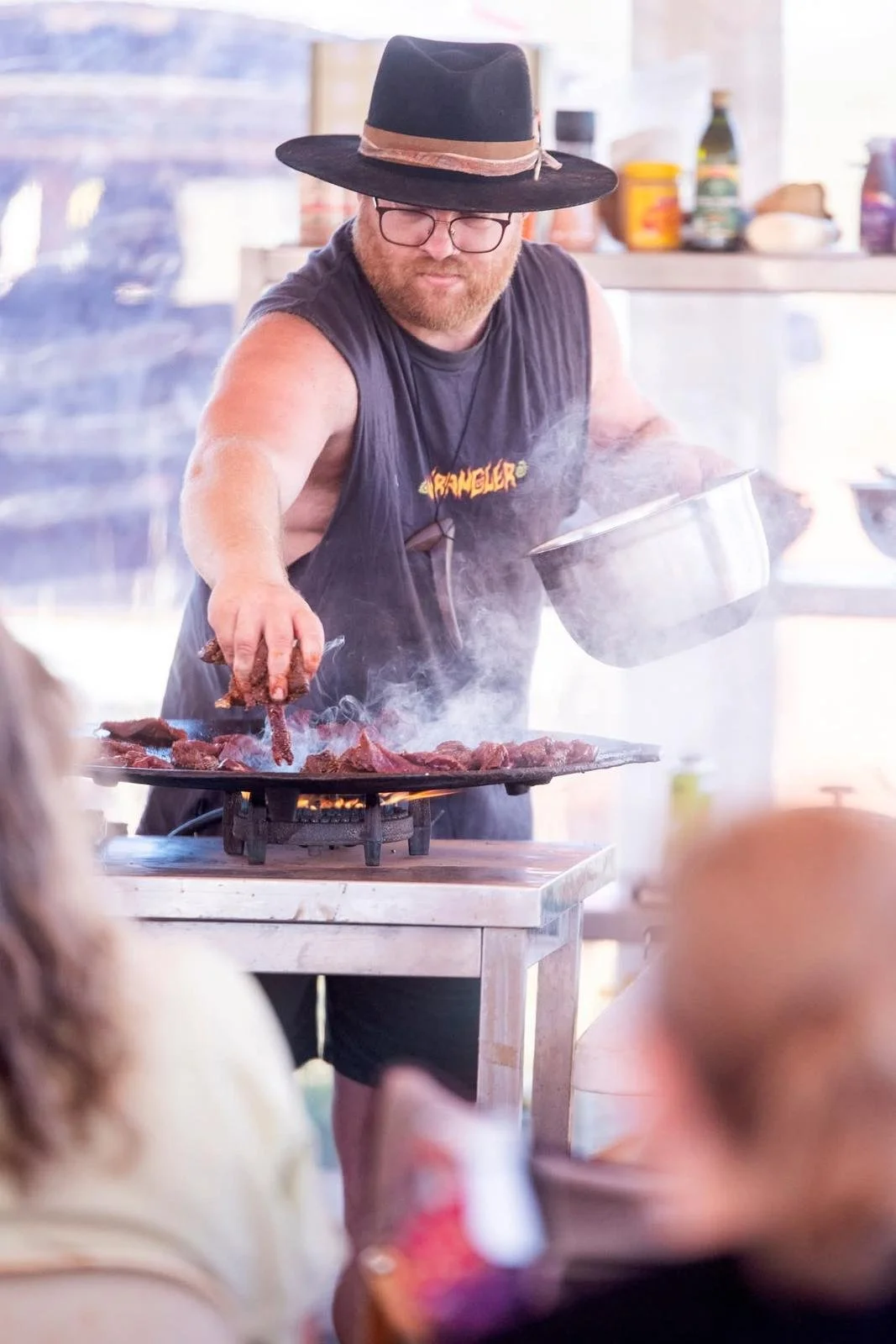 A man wearing a black hat, glasses, and a sleeveless shirt cooking meat on a flat grill with smoke rising. He is holding a pot in one hand and using tongs in the other to handle the meat, in a bright, casual kitchen or outdoor setting.