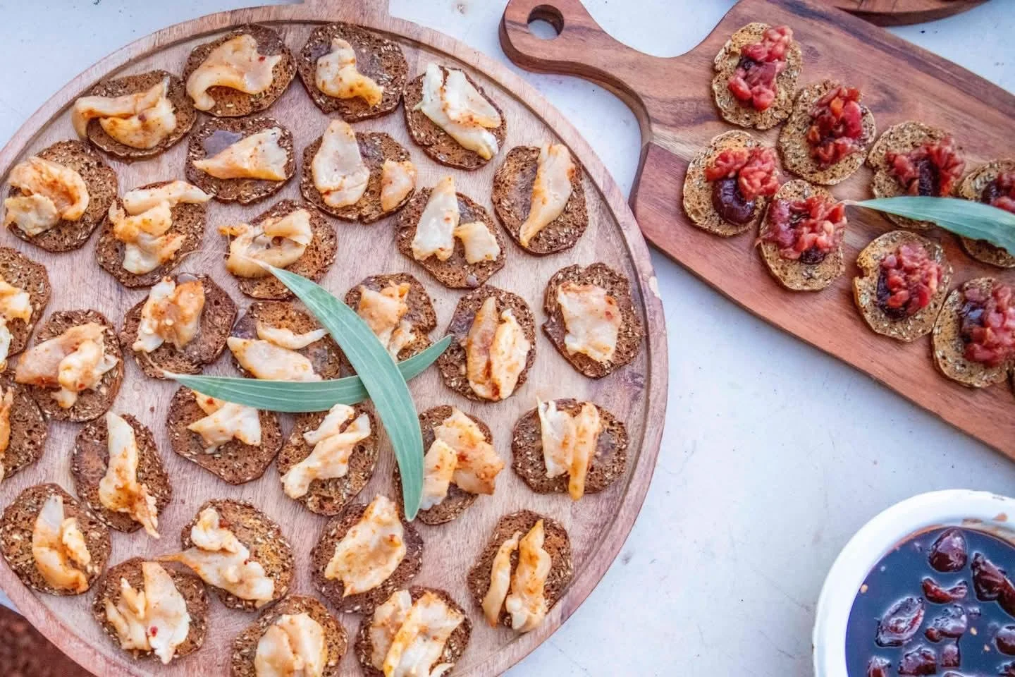 Assorted crackers topped with cheese, spread with toppings like tomato, onion, or possibly pâté, arranged on round and rectangular wooden serving boards, served with a bowl of dark-colored jam or chutney.