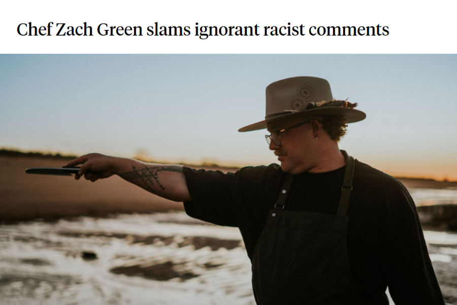 A man wearing a wide-brimmed hat, glasses, and a black apron, standing outdoors at sunset, pointing with his right arm extended toward the water.