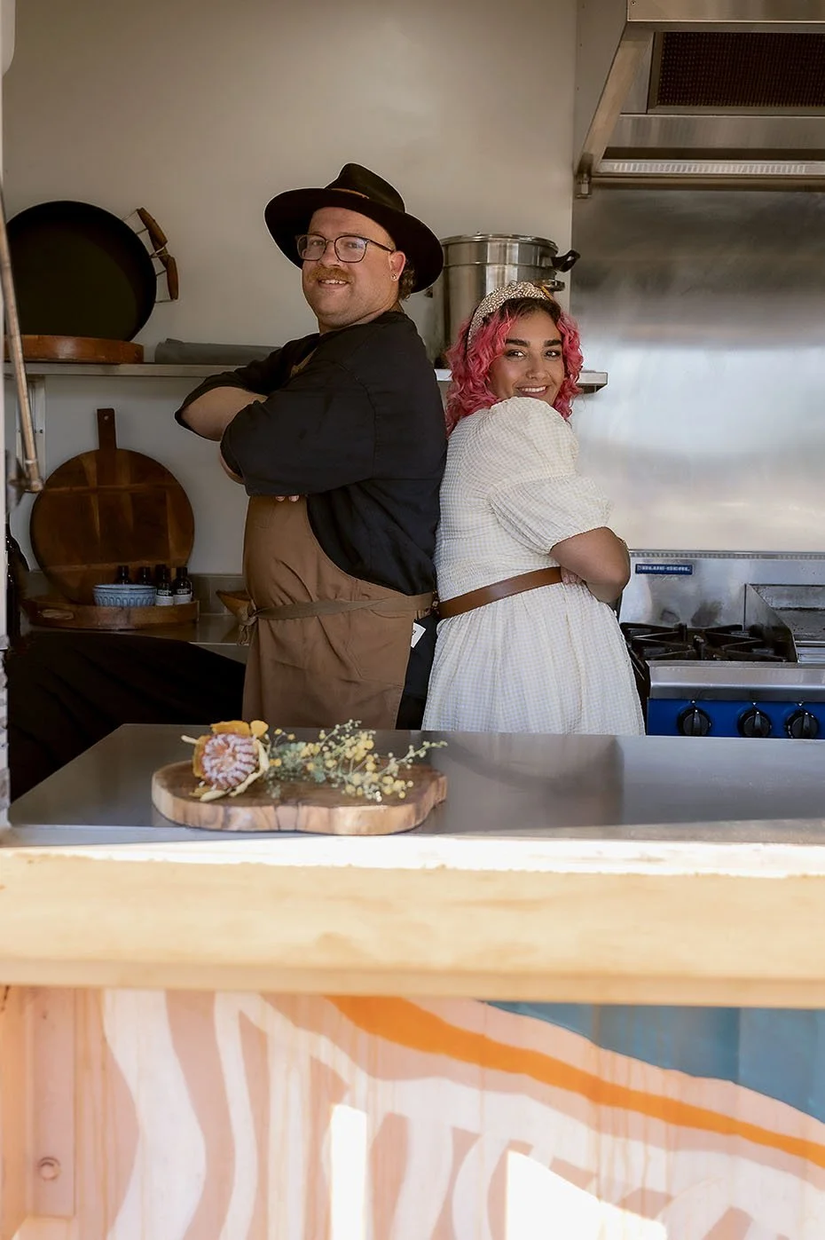 Two chefs, a man with glasses and a black hat, and a woman with pink curly hair and a white dress, standing back-to-back in a kitchen with arms crossed, smiling.