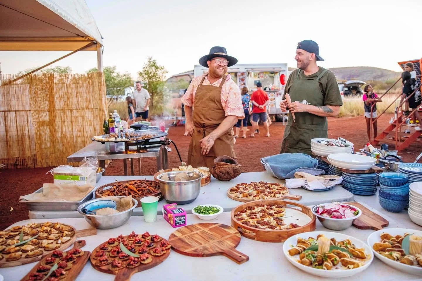 Outdoor food event with two smiling men in aprons and hats preparing and serving food on a table, surrounded by various dishes including pizza, pasta, and seafood, with a food truck and people in the background during daytime.