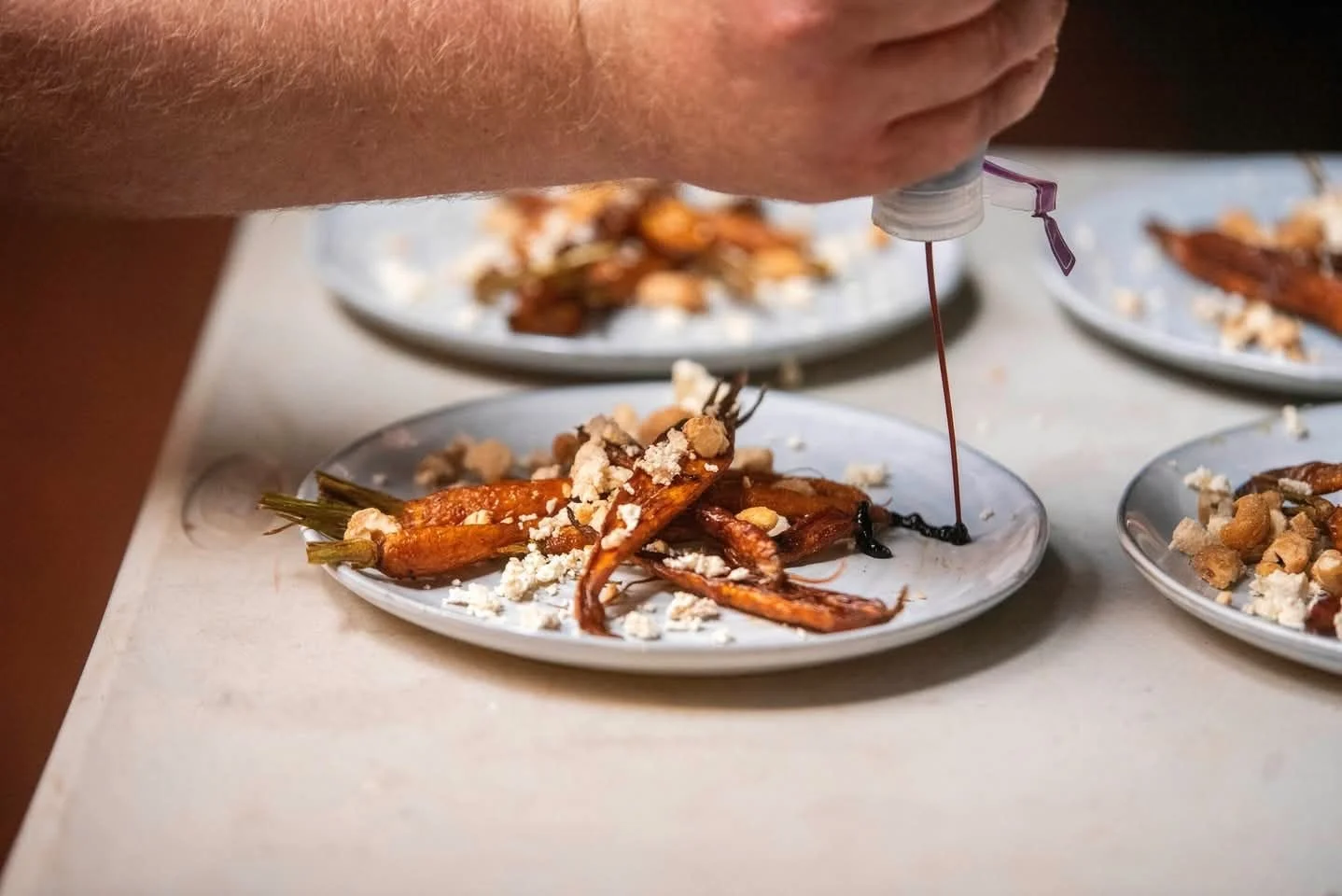 A chef uses a squeeze bottle to add sauce or oil to plated roasted carrots topped with crumbled cheese.