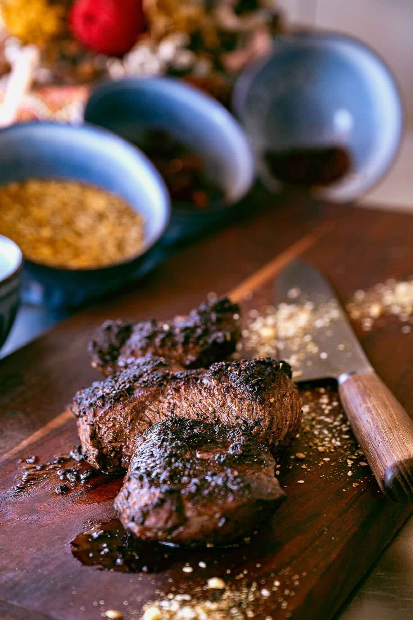Cooked steak on a wooden cutting board with a knife and scattered seasoning, with condiment bowls in the background.