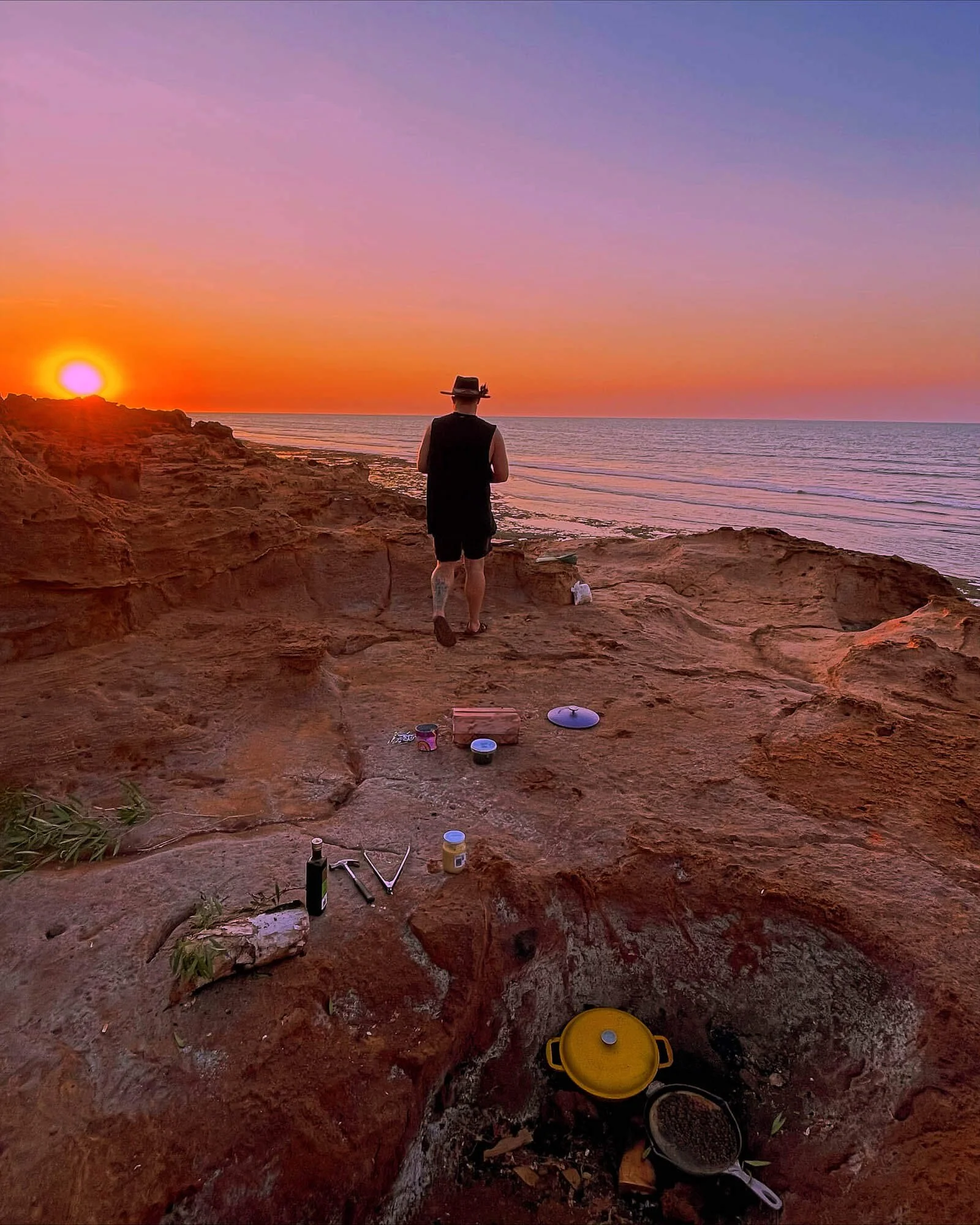 Person in a hat and sleeveless shirt standing on rocky beach at sunset, with cooking equipment and food items nearby.