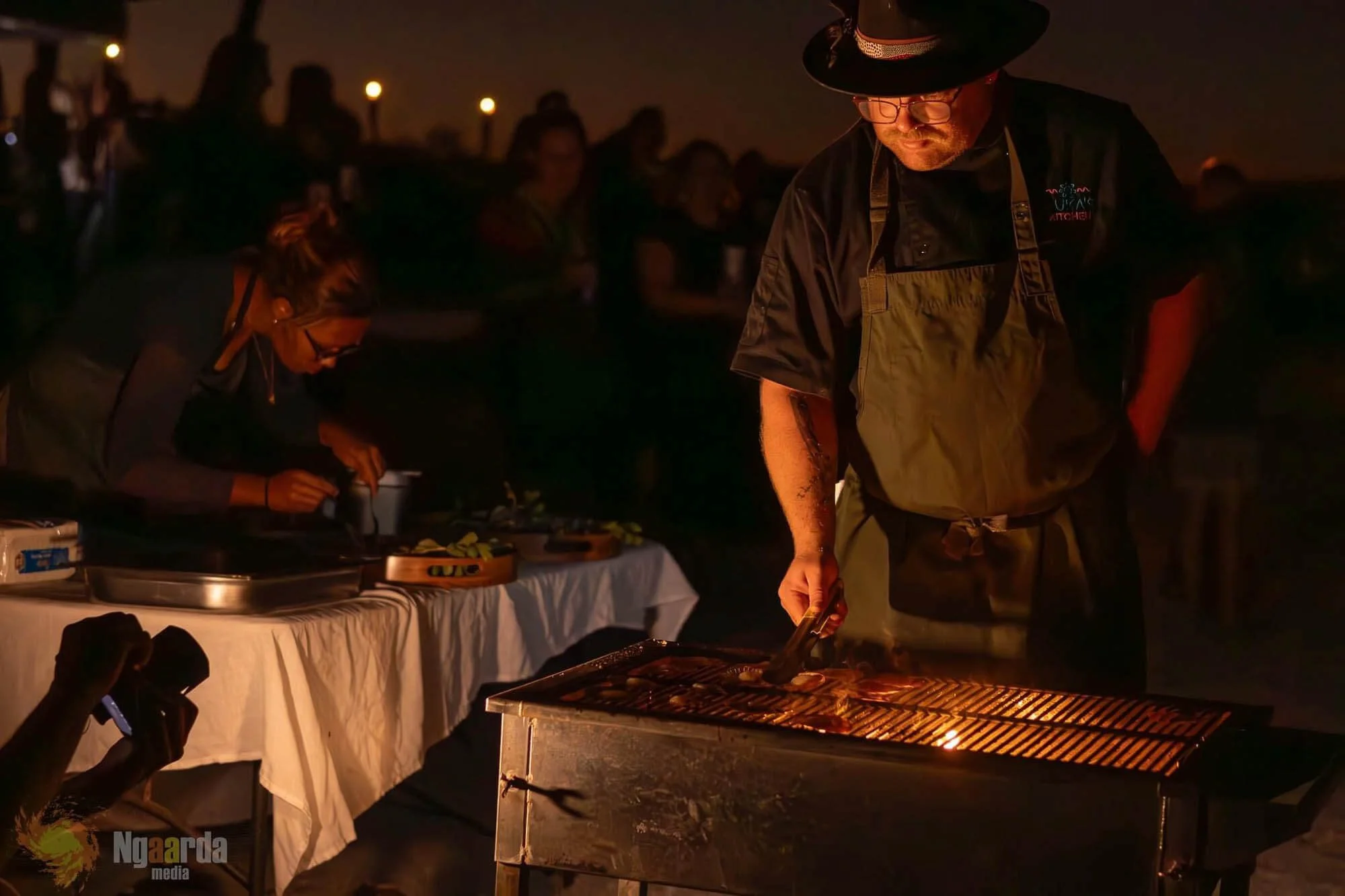 A man wearing a hat, glasses, black shirt, and apron grills food at night, with a table of food and people in the background.