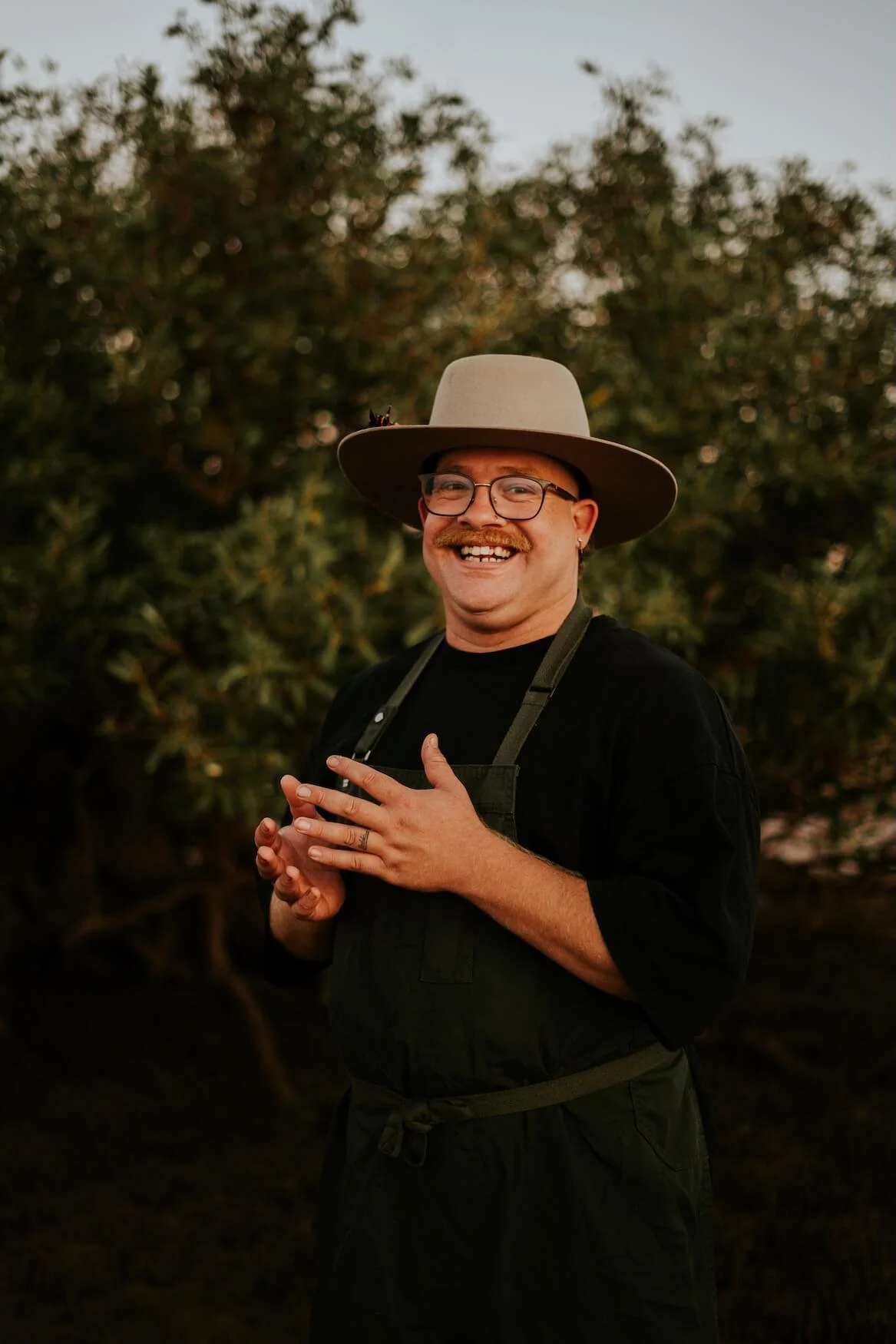 A smiling man with glasses, a mustache, and earrings, wearing a beige hat and black apron, standing outdoors with green foliage in the background.