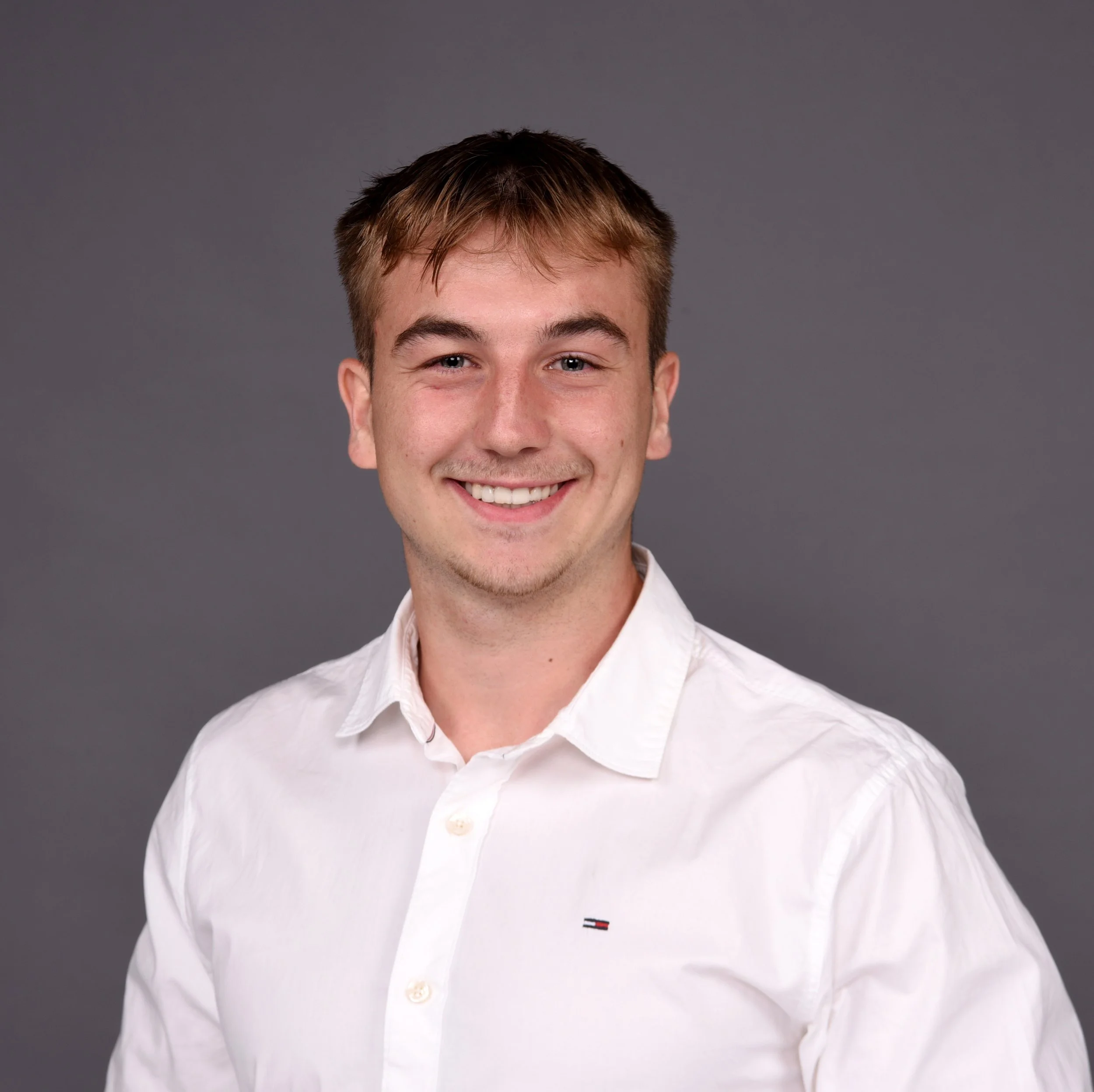 Porträt eines lachenden jungen Mannes mit braunen Haaren auf grauem Hintergrund.