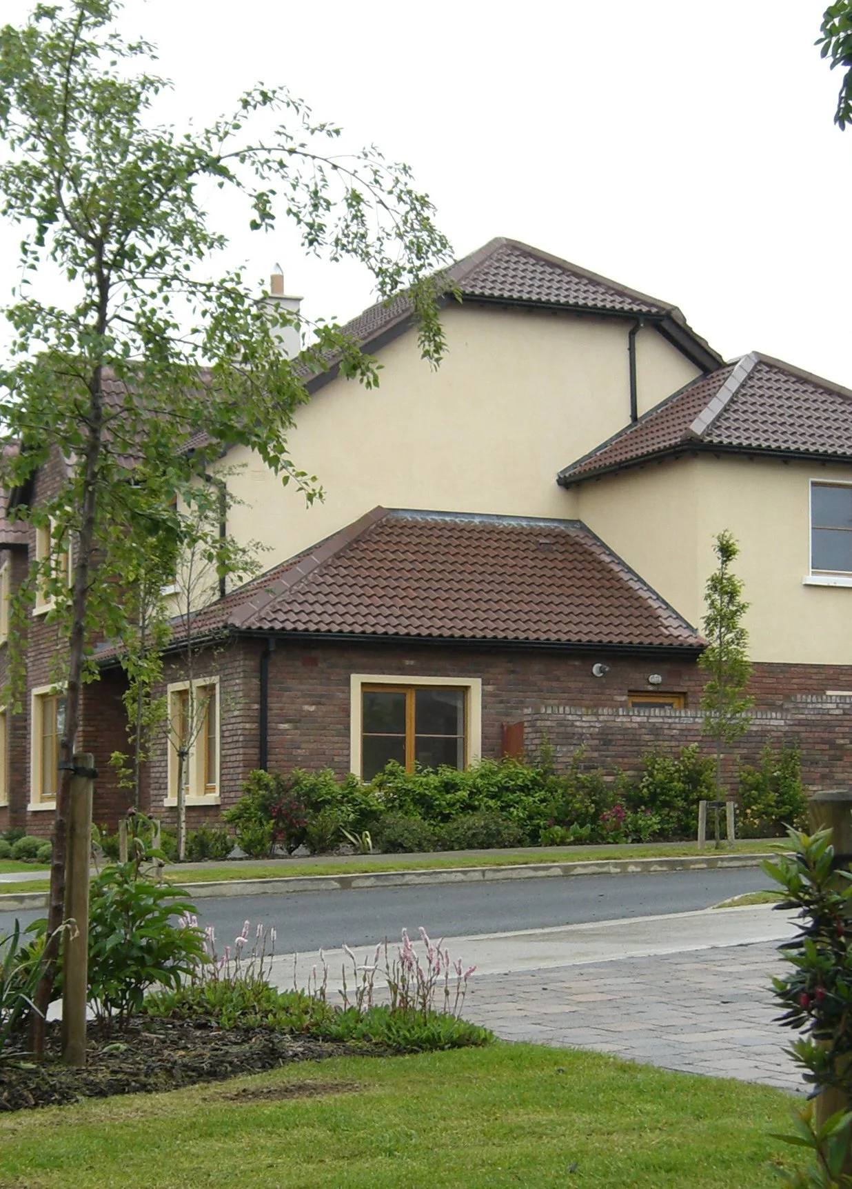 Residential house with brick and stucco exterior, brown tiled roof, surrounded by landscaped garden and trees.