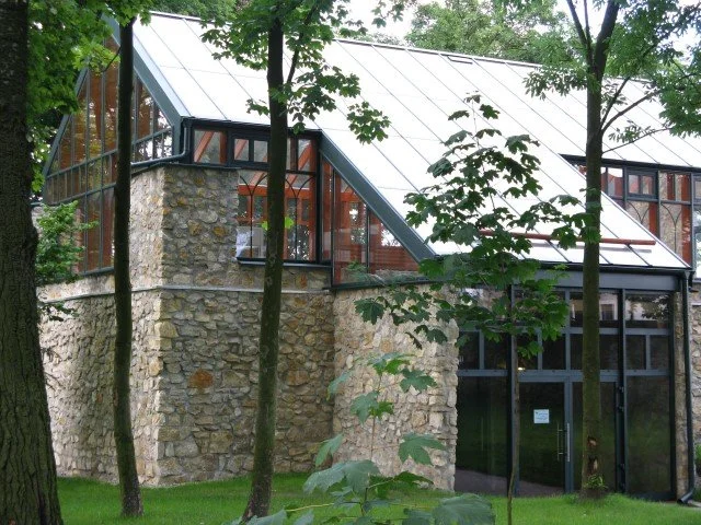 Stone and glass building with large windows and a sloped roof, surrounded by trees and greenery.