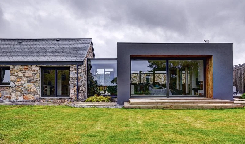 Modern house exterior with stone and dark gray facade, large glass windows, and a well-maintained lawn.
