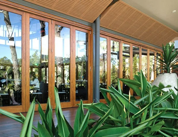 Glass folding doors with wooden frames leading to an outdoor patio with greenery and a ceiling with a slatted wood design.