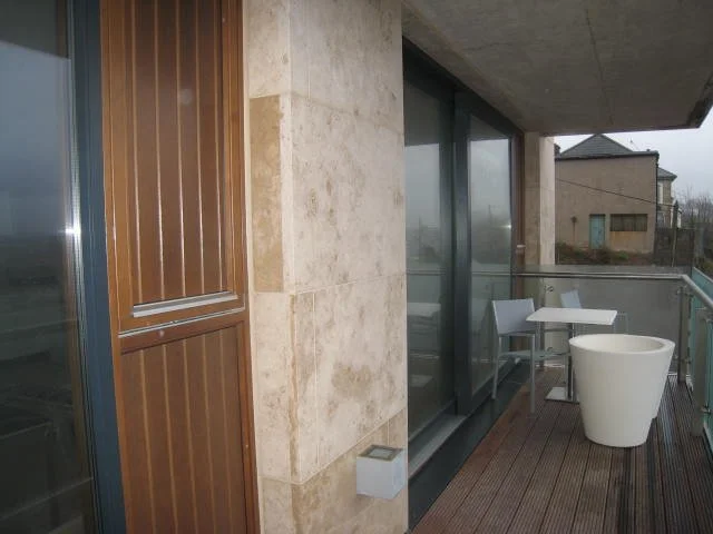 Modern balcony with glass railing, white table and chairs, and a large planter. The floor is wooden, and the building has a combination of stone and wood paneling.