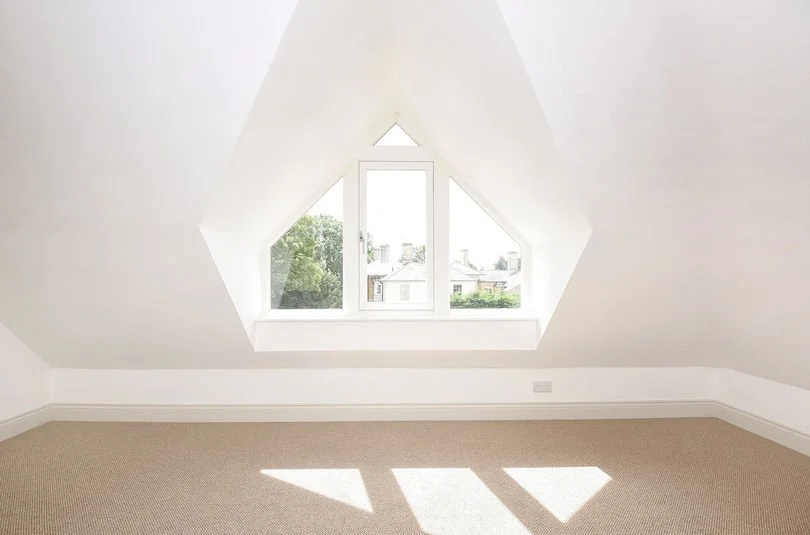 Bright attic room with triangular window, beige carpet, and white walls.