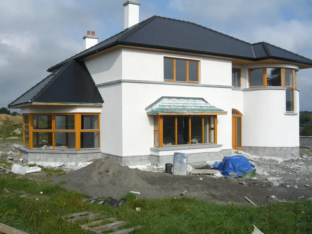 Two-story white house under construction with black roof and wooden framed windows, surrounded by construction debris and materials.