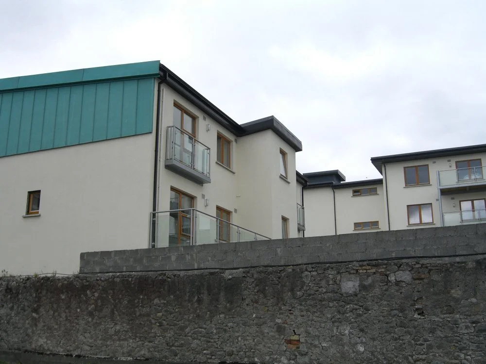 Modern residential building with multiple balconies, white walls, and teal roof panels against a cloudy sky.