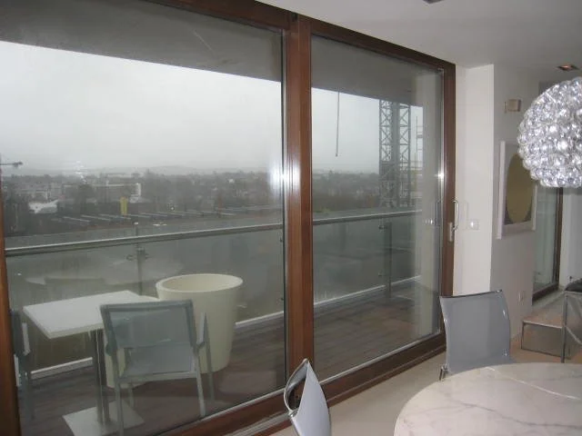 Interior view of a modern room with large sliding glass doors leading to a balcony, which includes patio furniture. The balcony overlooks a cityscape on a foggy day. Inside, there's a round marble table with gray chairs and a decorative light fixture