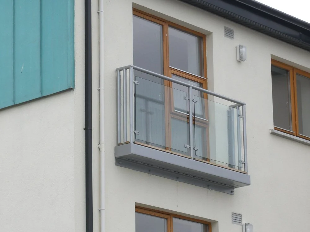 Modern glass balcony on a building with light stucco walls and wooden-framed windows.