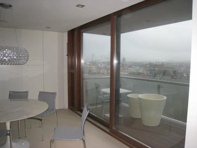 Modern dining area with round table, gray chairs, a large window with a city view, and a balcony with planters.