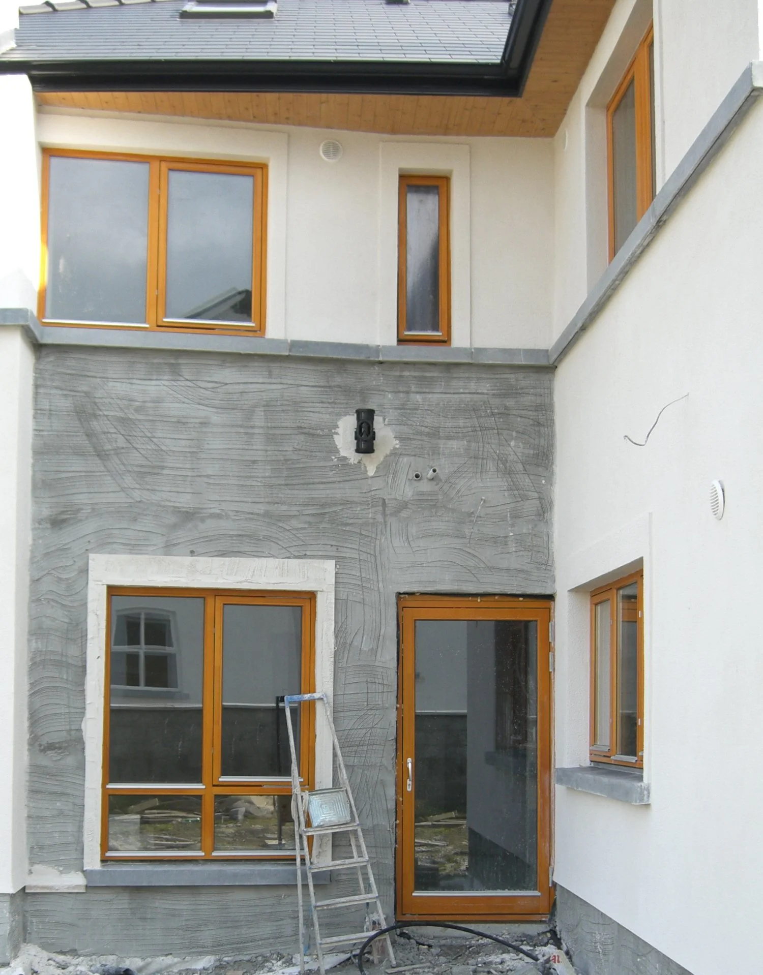 Two-story house exterior with white walls and wooden-framed windows under construction, featuring a ladder leaning against the wall and an unfinished plaster section.