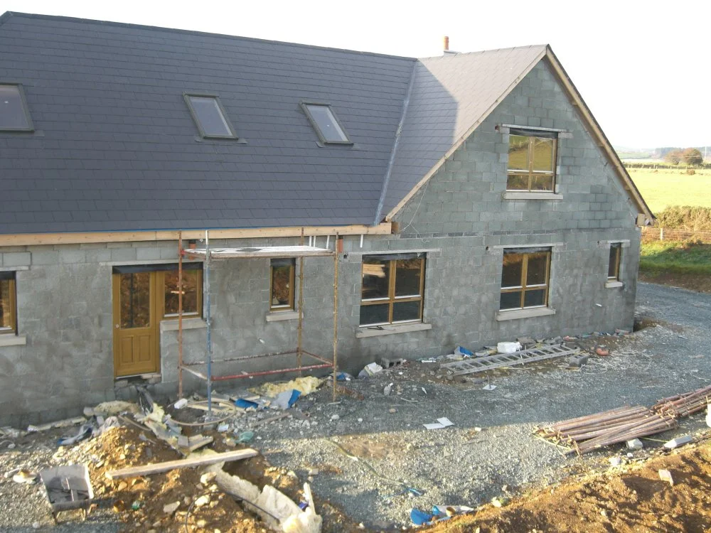 Partially constructed stone house with scaffolding, several windows, and a steep roof. Building materials and debris are scattered around the site.