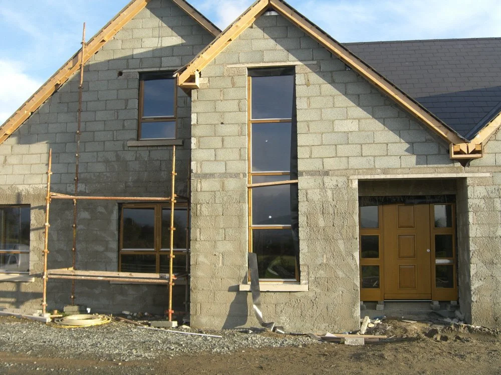 House under construction with cinder block walls, wooden roof framing, scaffolding, wooden front door, and partially completed windows.