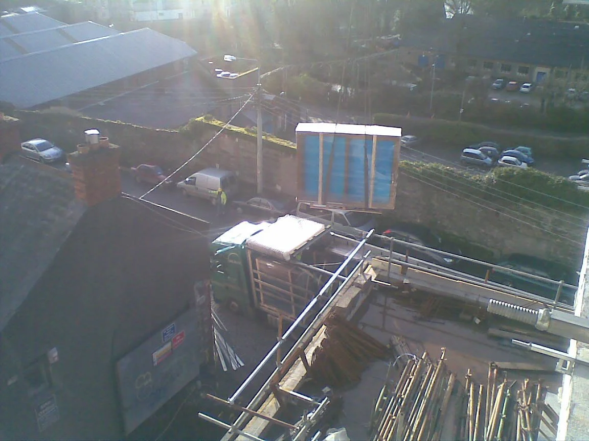 Construction site with a crane lifting a large box over a building, adjacent to parked cars and a road, under sunny conditions.