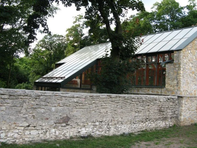 Stone building with glass roof and wooden accents surrounded by trees and a stone wall in a garden setting.