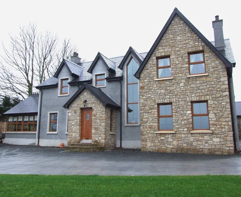 A large, two-story house with a mix of stone and stucco exterior, featuring multiple gabled roofs and wooden-framed windows. The front entrance has a wooden door and a hanging lantern. The house is set against a yard with trees in the background.