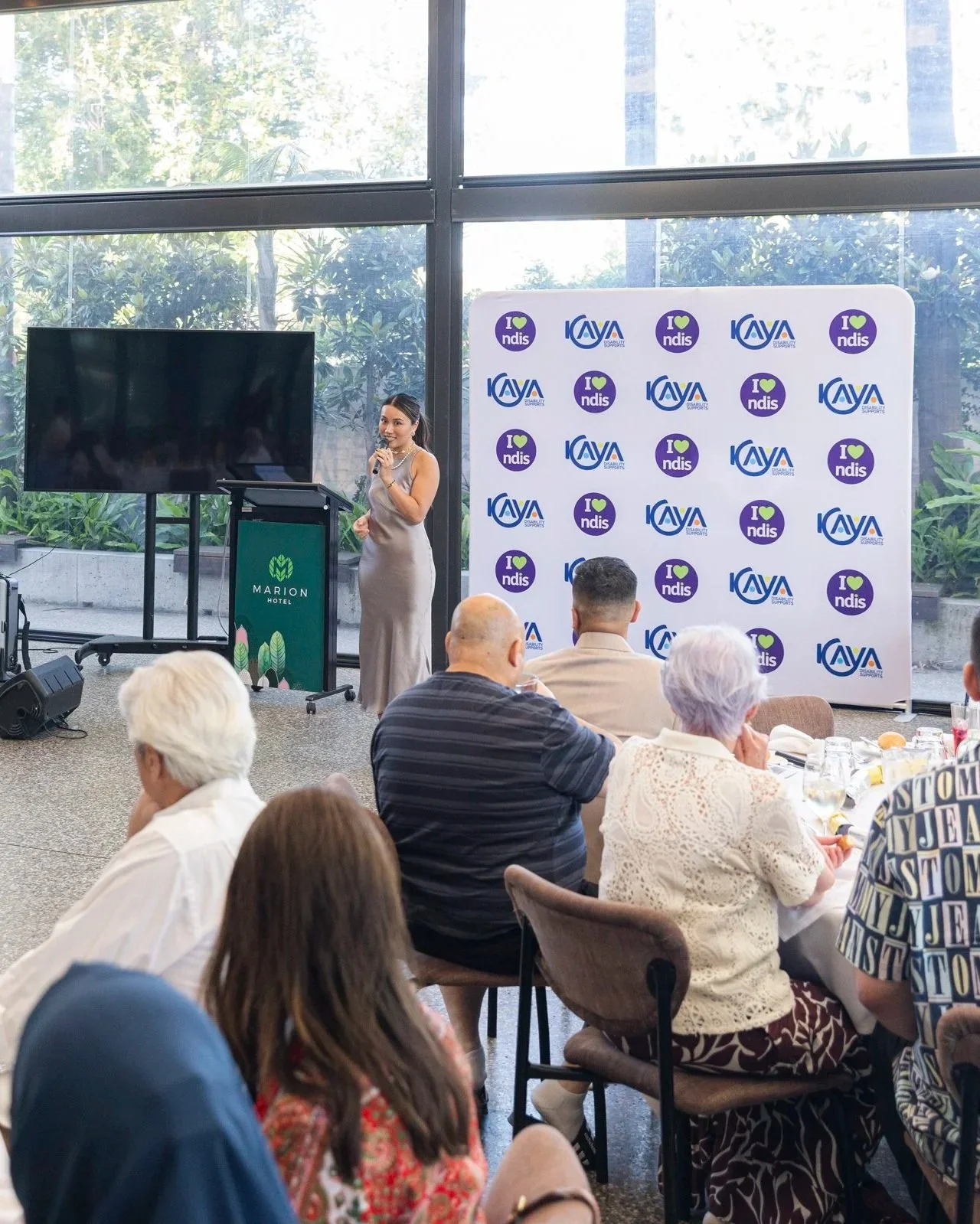 A woman in a silver dress standing on a stage with a microphone, speaking to an audience seated at tables in a conference room with large windows and greenery outside.