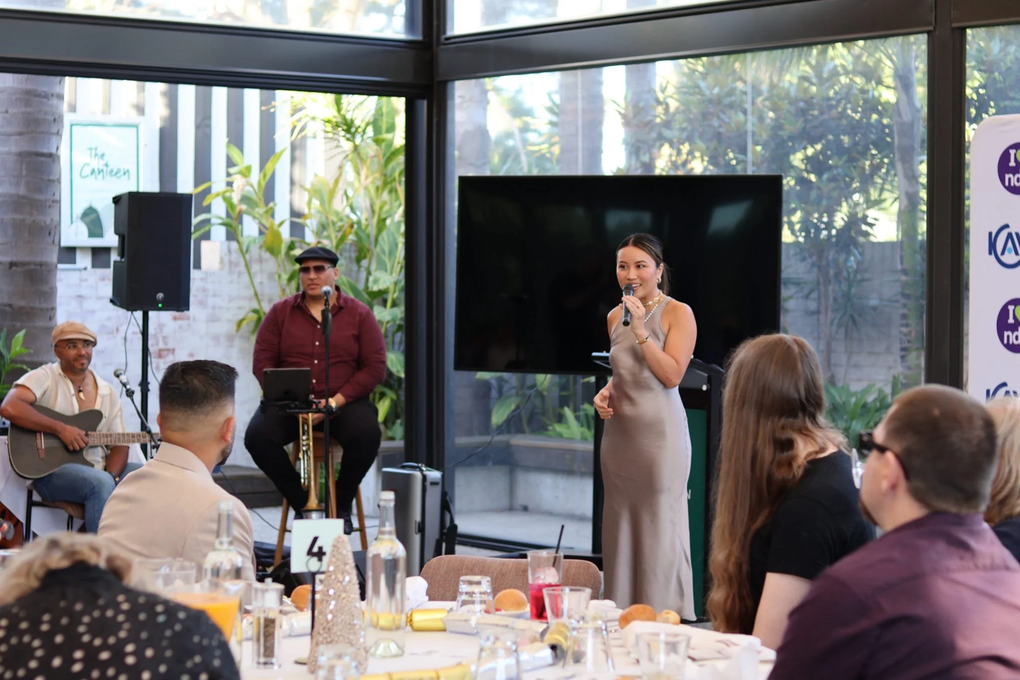 A woman in a beige gown giving a speech or performance at a formal event, with musicians playing instruments behind her and an audience seated at tables in a brightly lit venue with large windows and greenery outside.