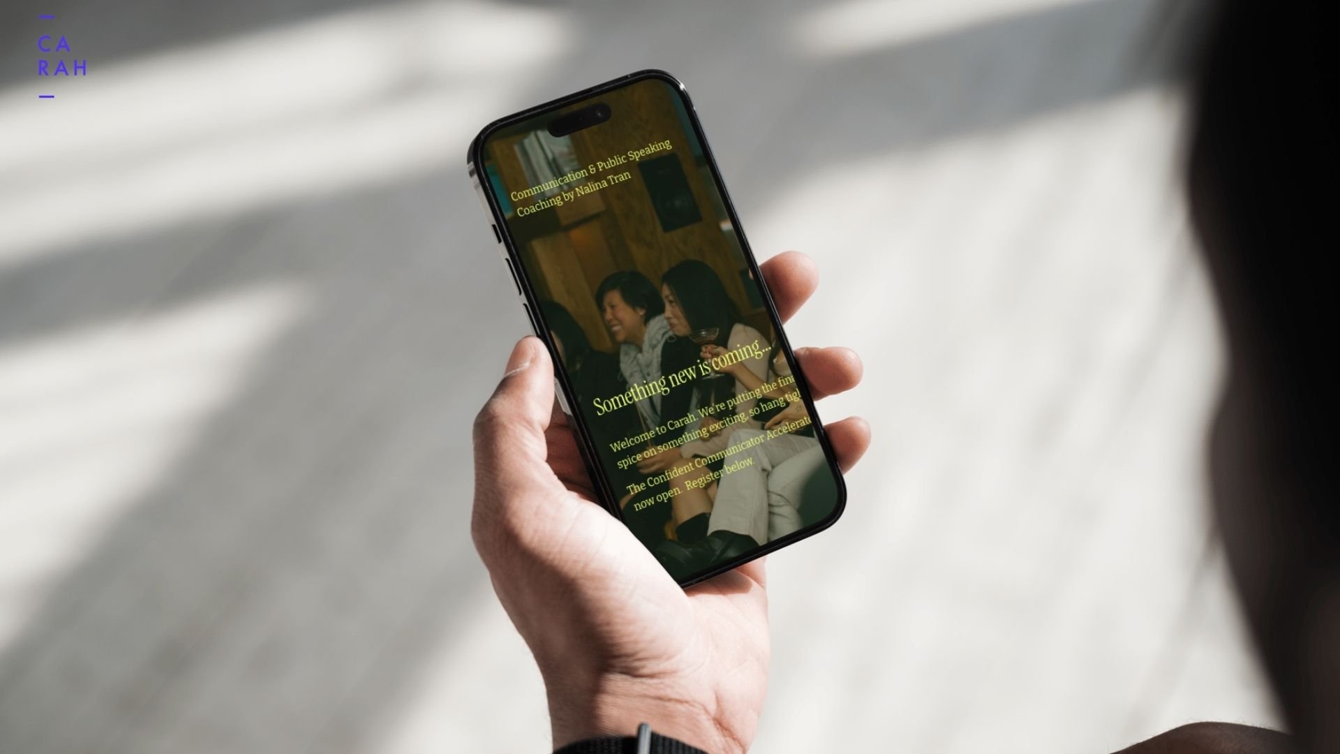 A hand holding a smartphone with a promotional screen for communication and public speaking coaching, showing a group of people at a social gathering with text overlay about upcoming events.