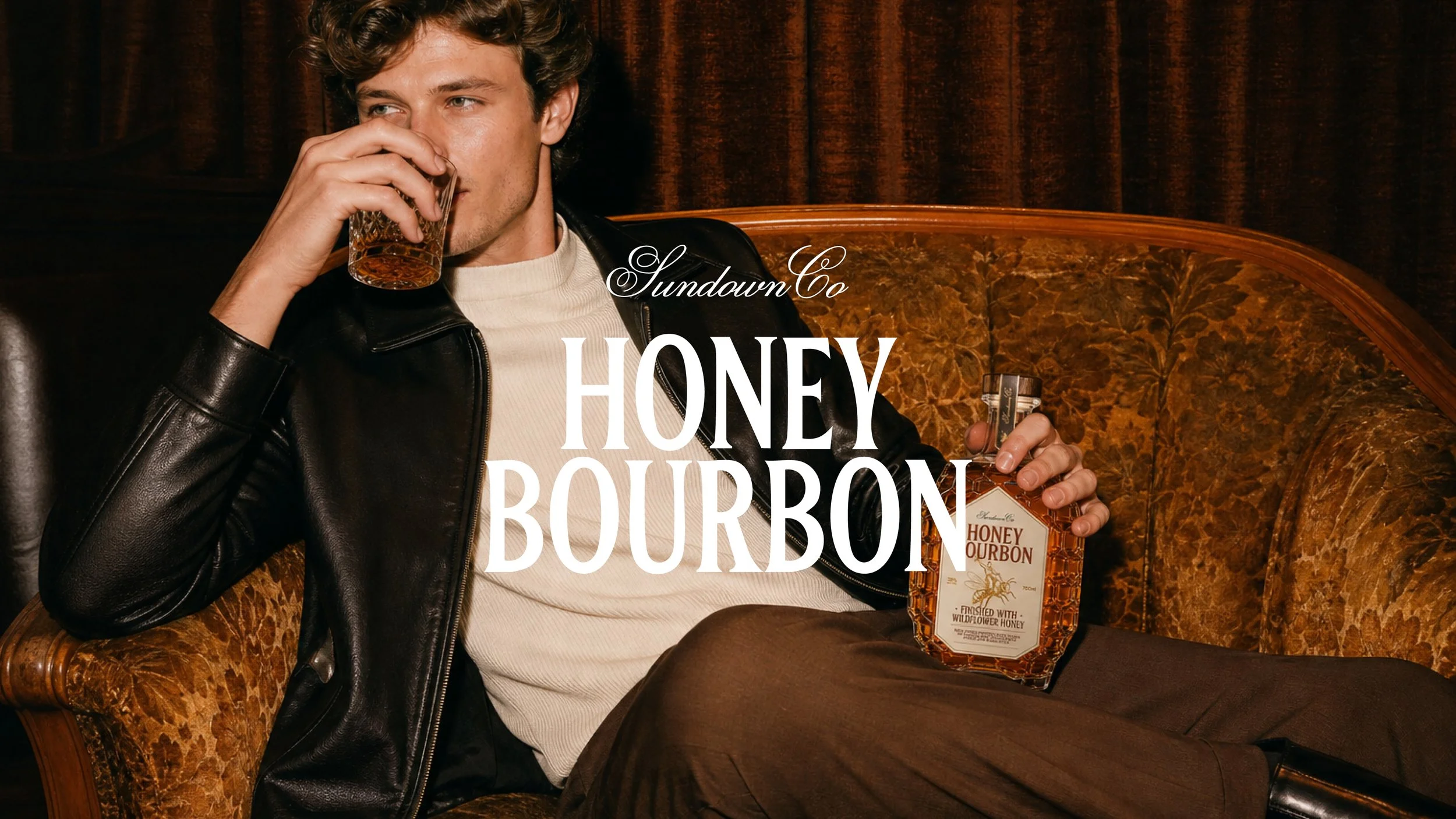 A young man with wavy hair sitting on a vintage sofa, drinking whiskey from a glass, with a bottle of Honey Bourbon whiskey on his lap, dressed in a white shirt and black leather jacket, in a dimly lit room with dark curtains.
