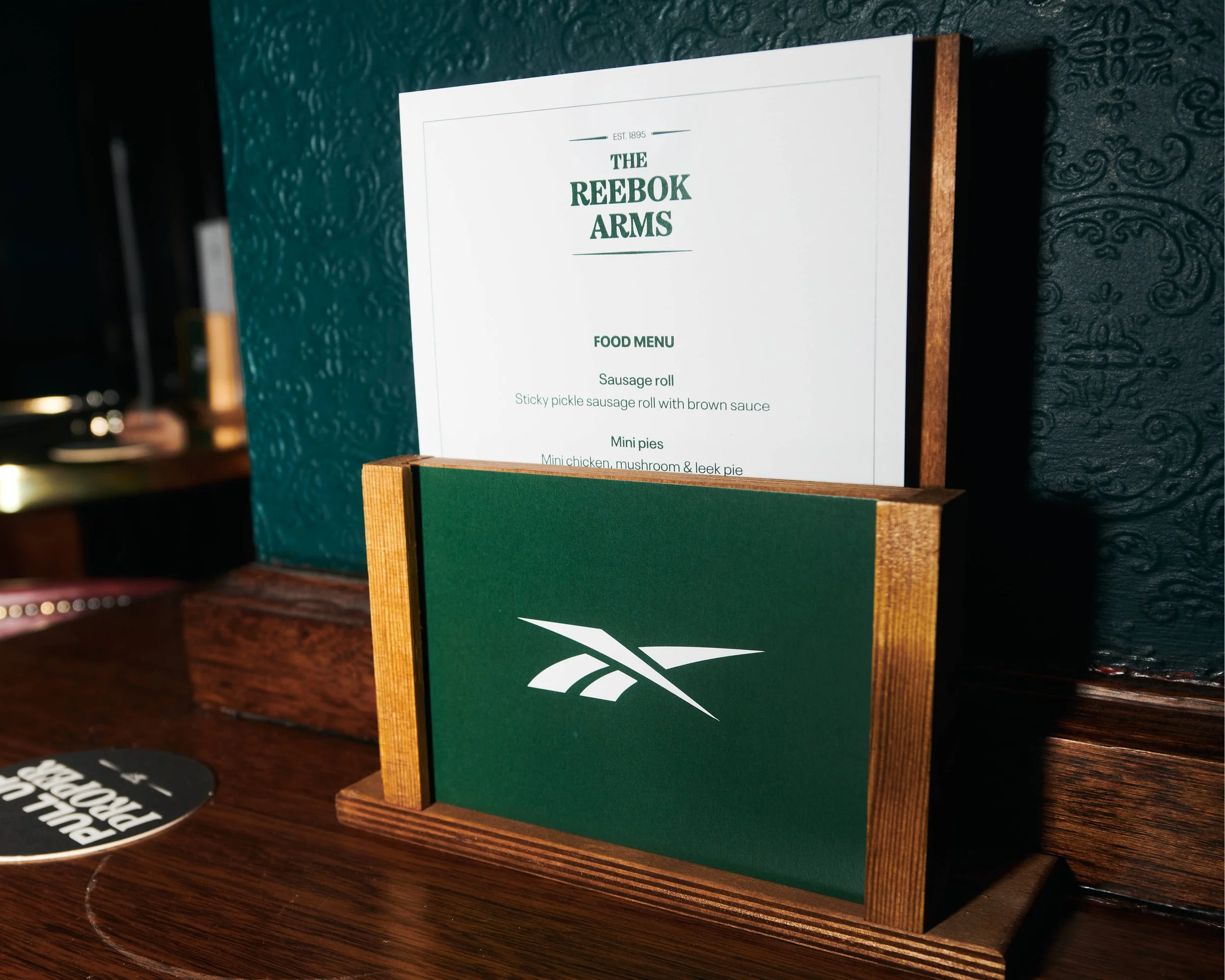 Menu sign for The Reebok Arms pub on a wooden stand, featuring food options such as sausage roll and mini pies, against a textured dark wall background.