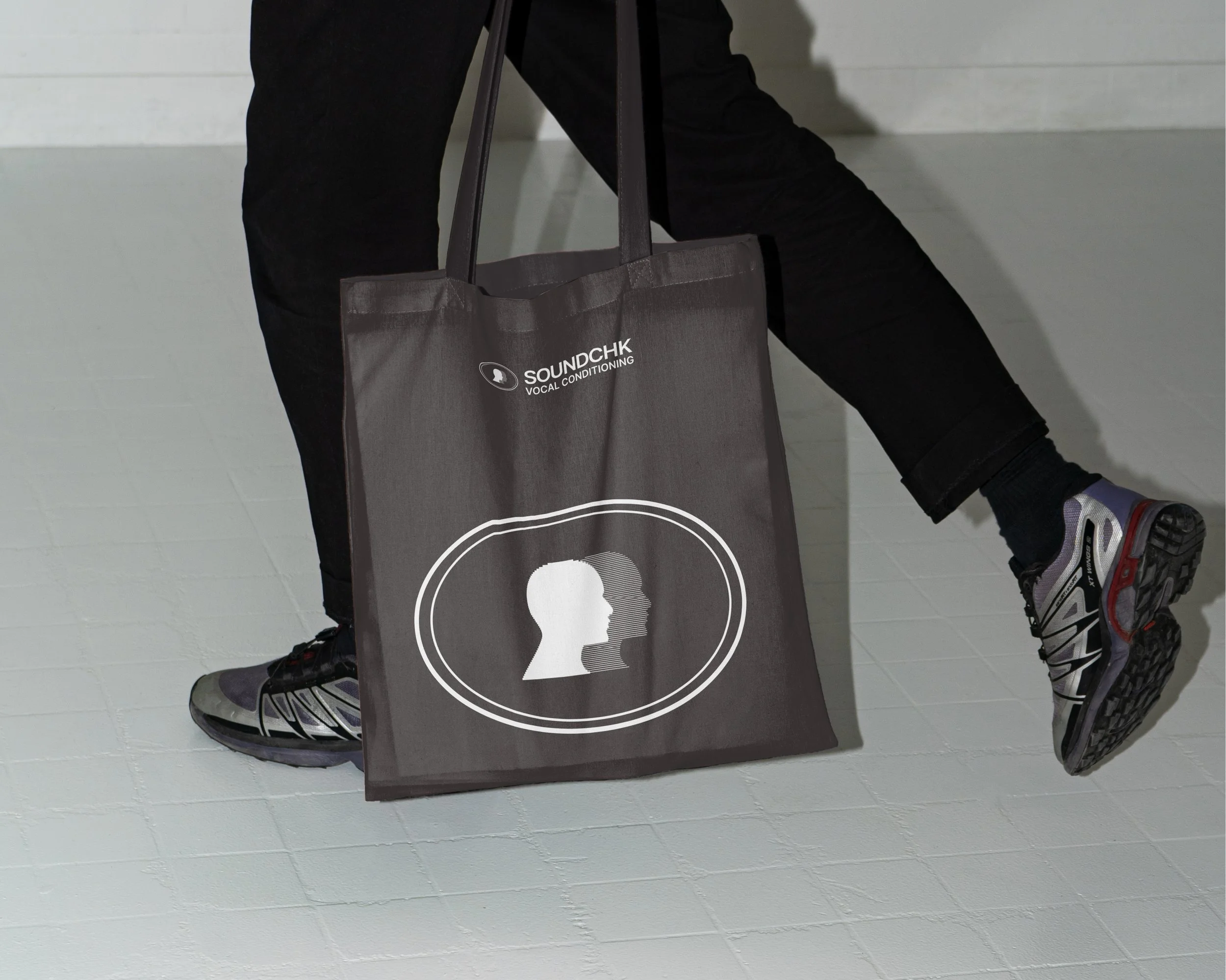 A person carrying a black tote bag with the logo and text for SoundChak Vocal Conditioning, wearing sneakers and black pants, walking on a white tiled floor.