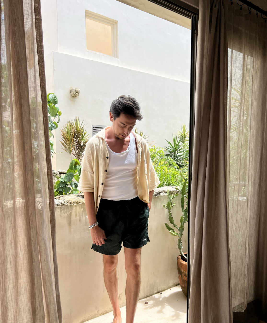 A man standing outside on a balcony, looking down, dressed in a white tank top, a light unbuttoned short-sleeve shirt, and black shorts, surrounded by green plants and a white building wall.