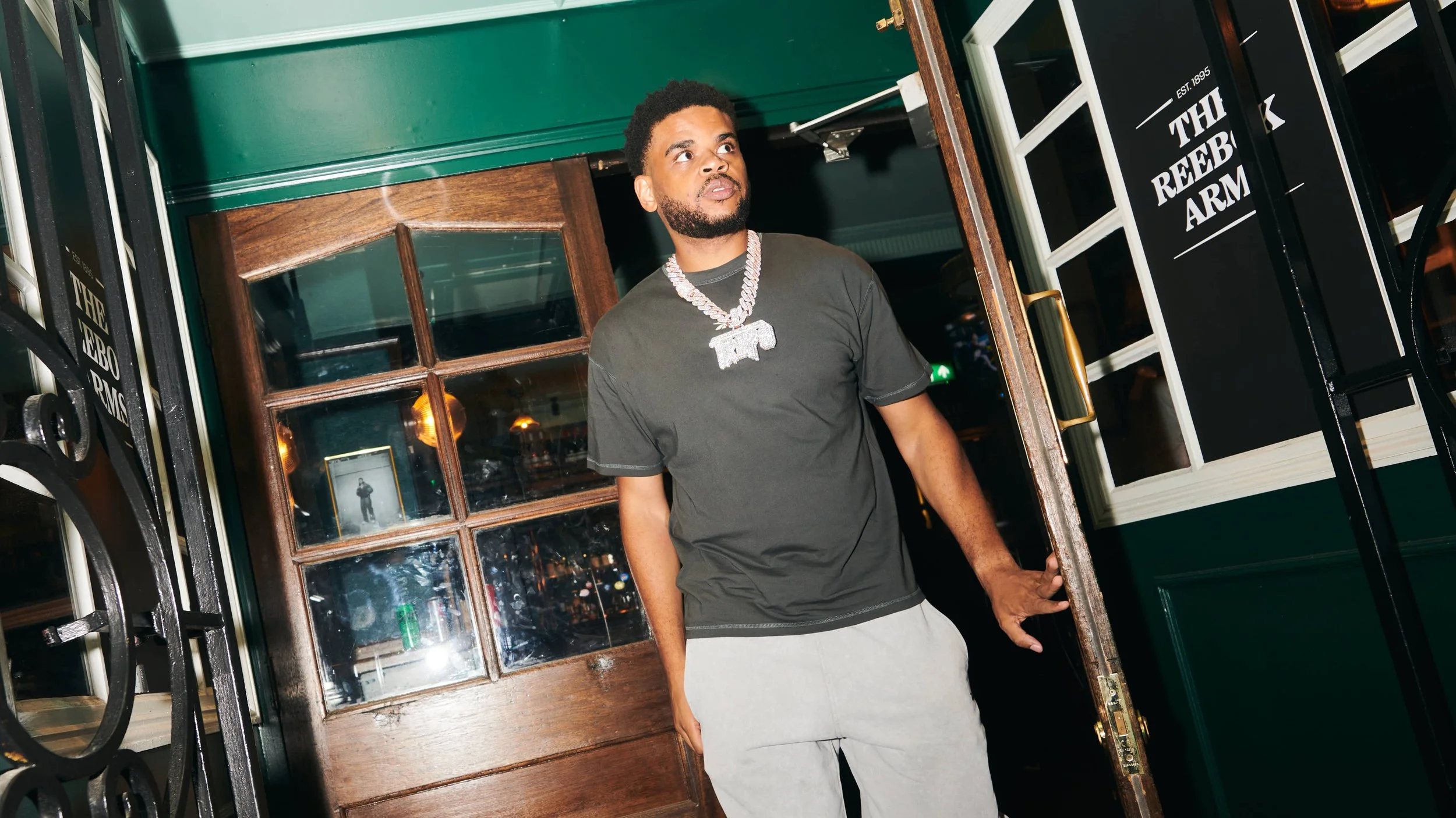 A young man with a beard wearing a black t-shirt and gray sweatpants is opening a restaurant door. He is wearing a large diamond chain and looking upward.