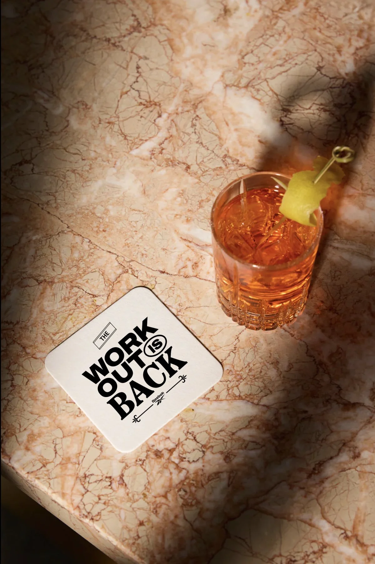 A drink with ice and a lemon wedge garnish on a pink and beige marble table. A white coaster with bold black text reads "The work outs is back."