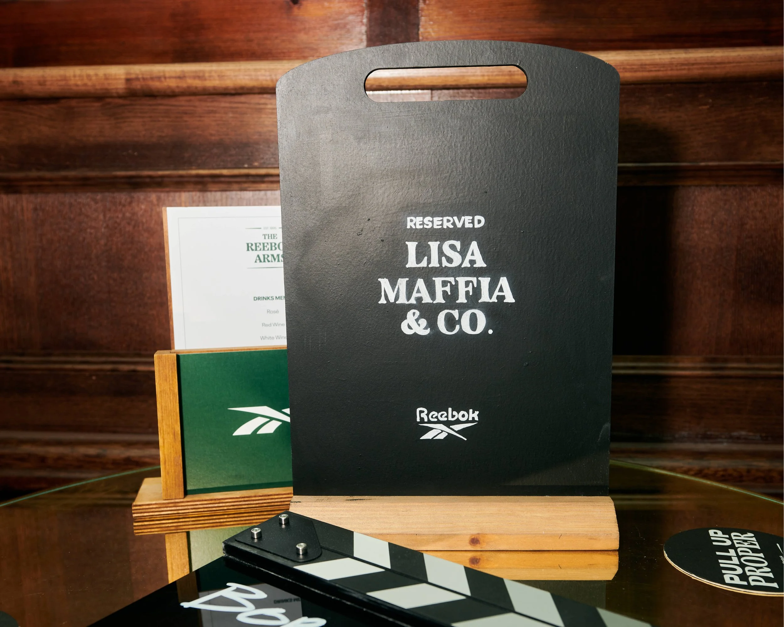 A black reserved sign with white text displaying the name "Lisa Maffa" and the logo of Reebok at the bottom, placed on a wooden surface with wooden panels in the background.