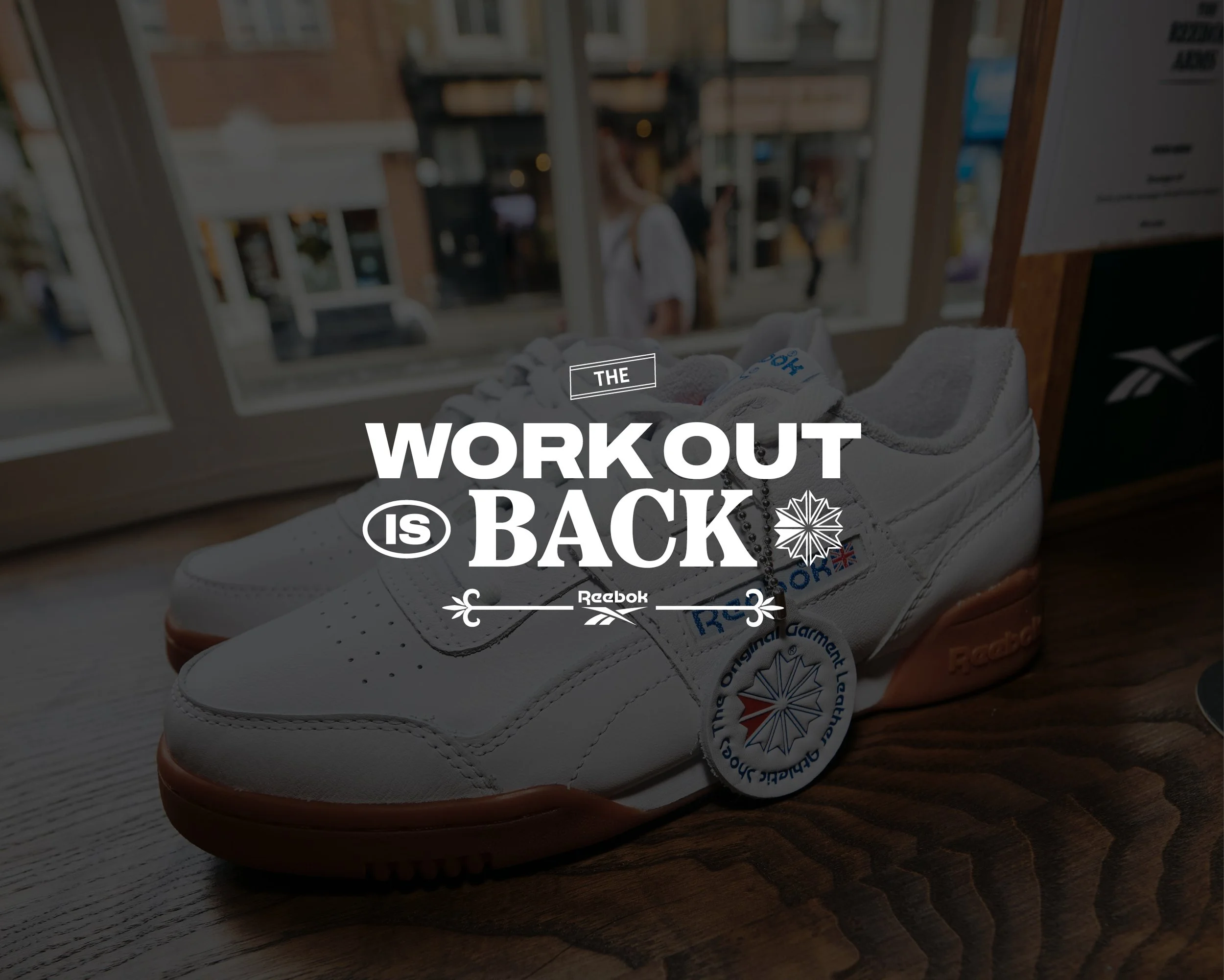 A pair of white athletic shoes with Reebok branding and a tag, resting on a wooden surface inside a store, with a storefront window showing people outside.