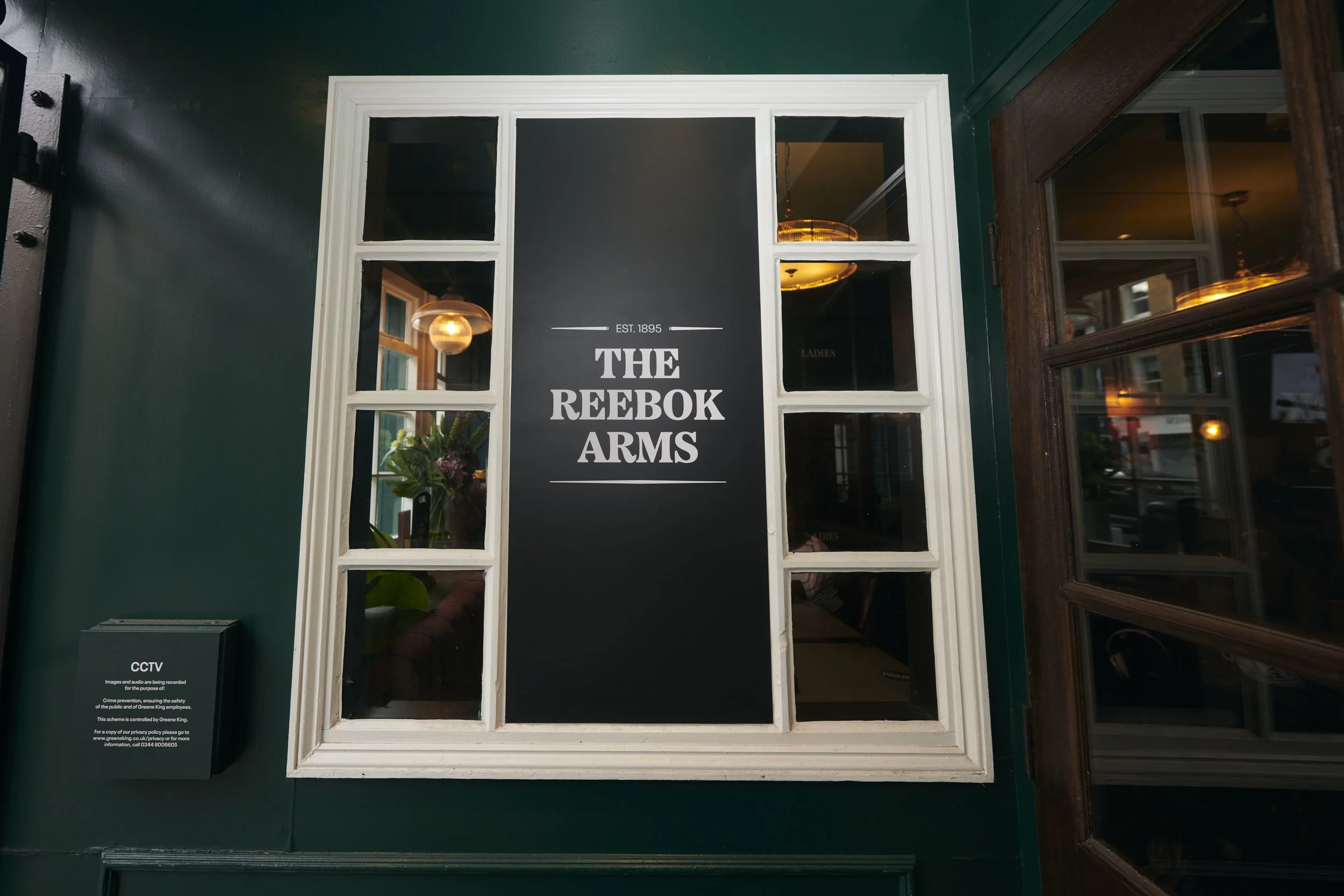 Storefront window with a black sign that reads "The Reebok Arms" with establishment year 1895, framed by white window panes, with interior lighting visible through the window panes.