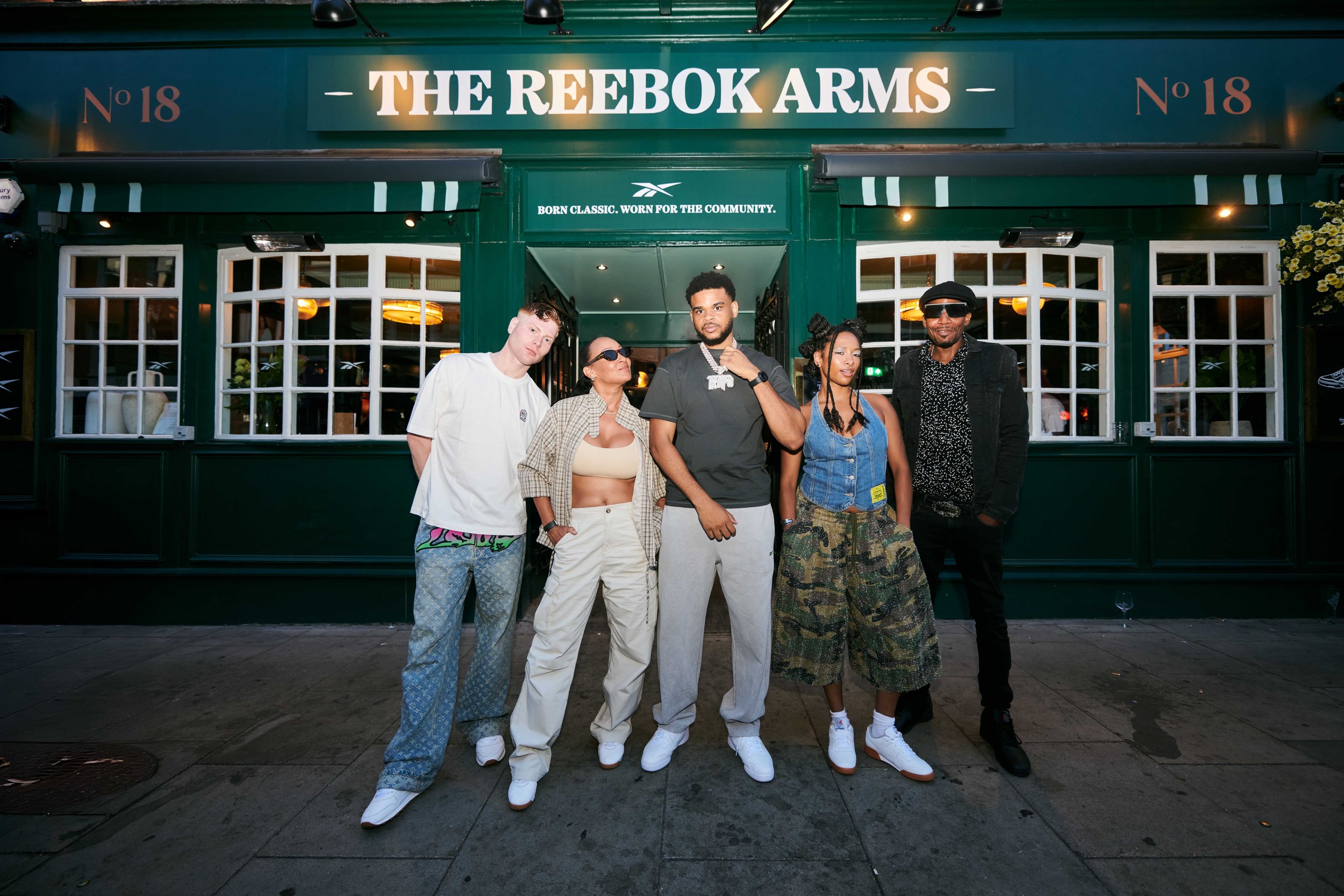 Group of five diverse people standing in front of a green storefront with the sign 'The Reebok Arms' and the number '18'. They are dressed in casual, trendy clothing, some wearing sunglasses, and posing confidently.