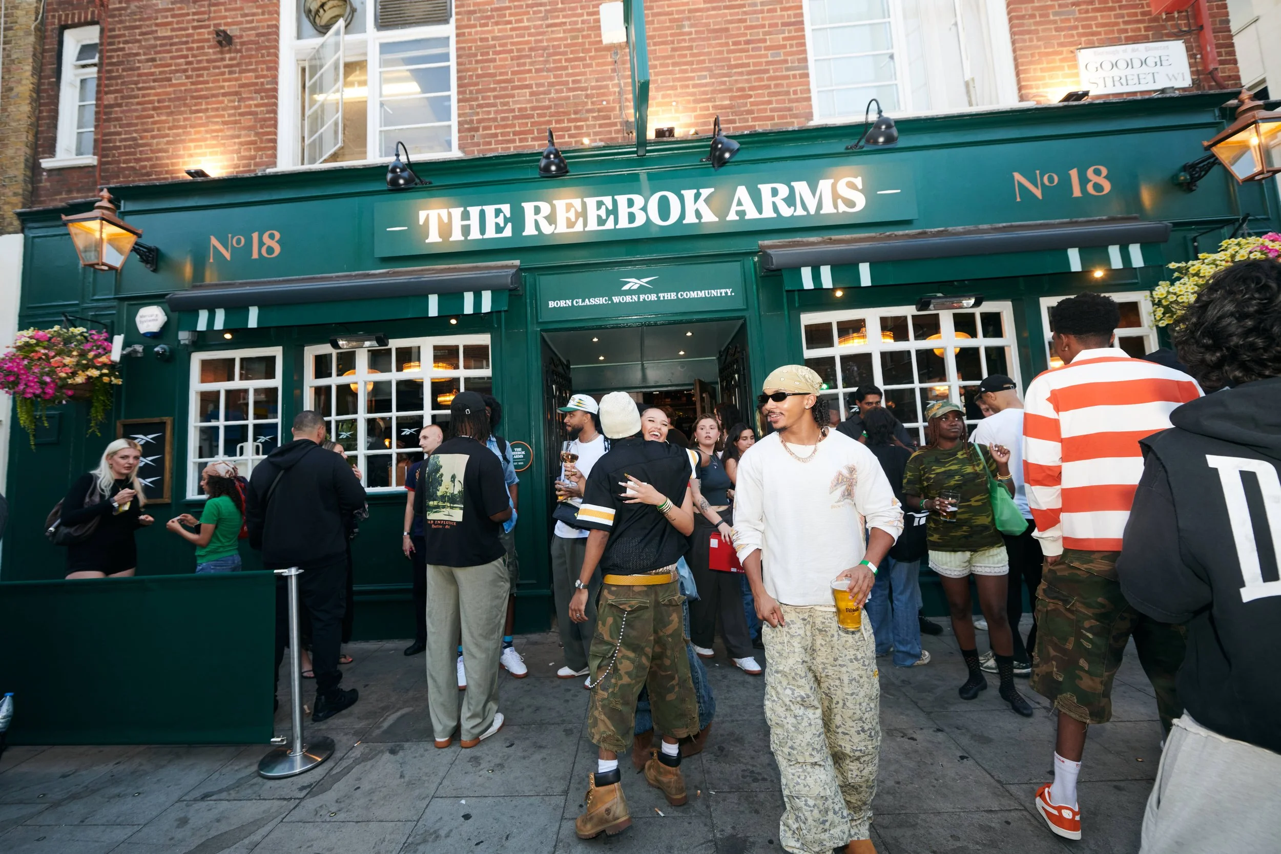 People gathered outside a pub called The Reebok Arms, with some holding drinks and engaging in conversation, in what appears to be a lively social scene.