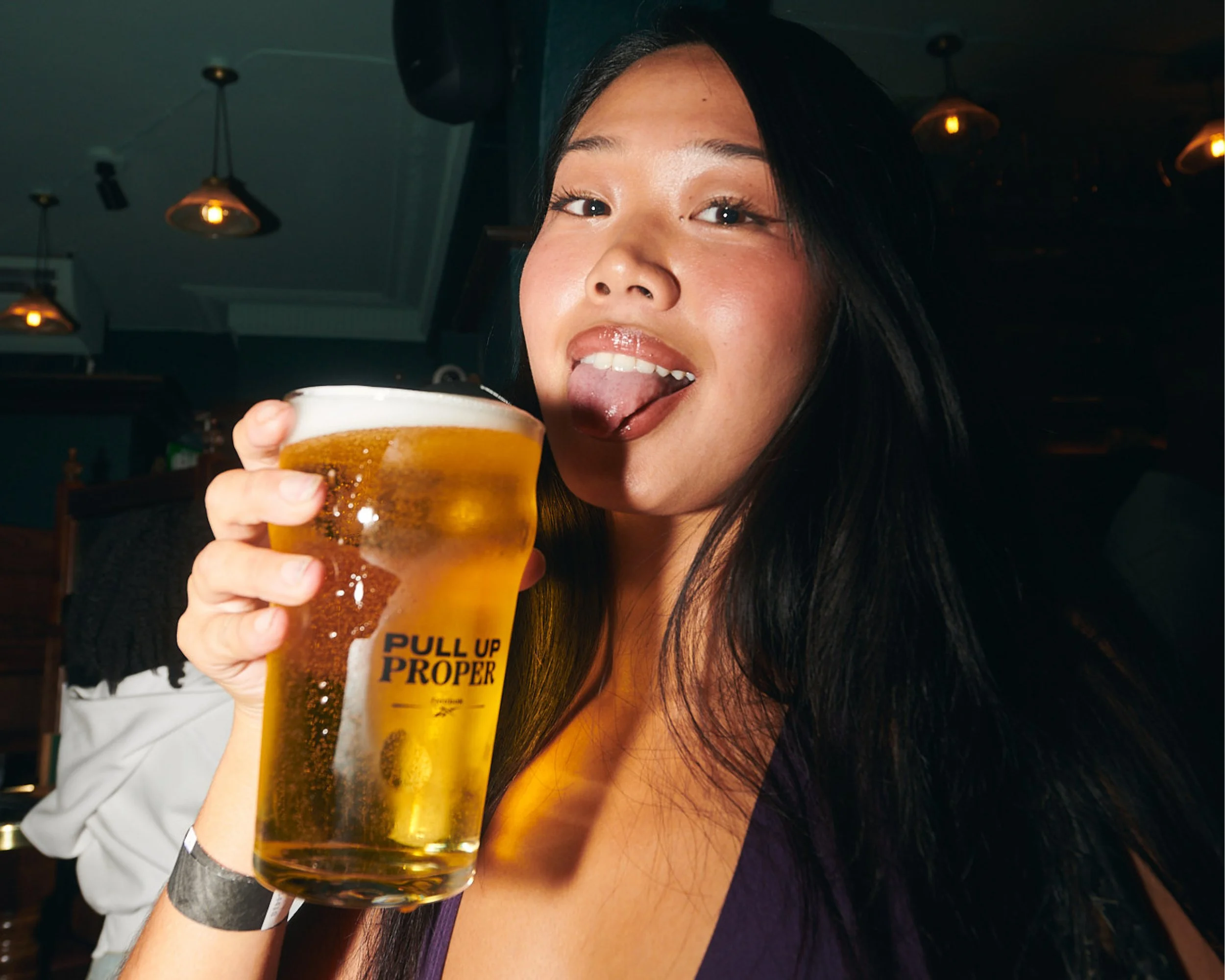 Close-up of a young woman with long dark hair smiling and holding a glass of beer at a bar or pub, with warm lighting and ceiling lights in the background.