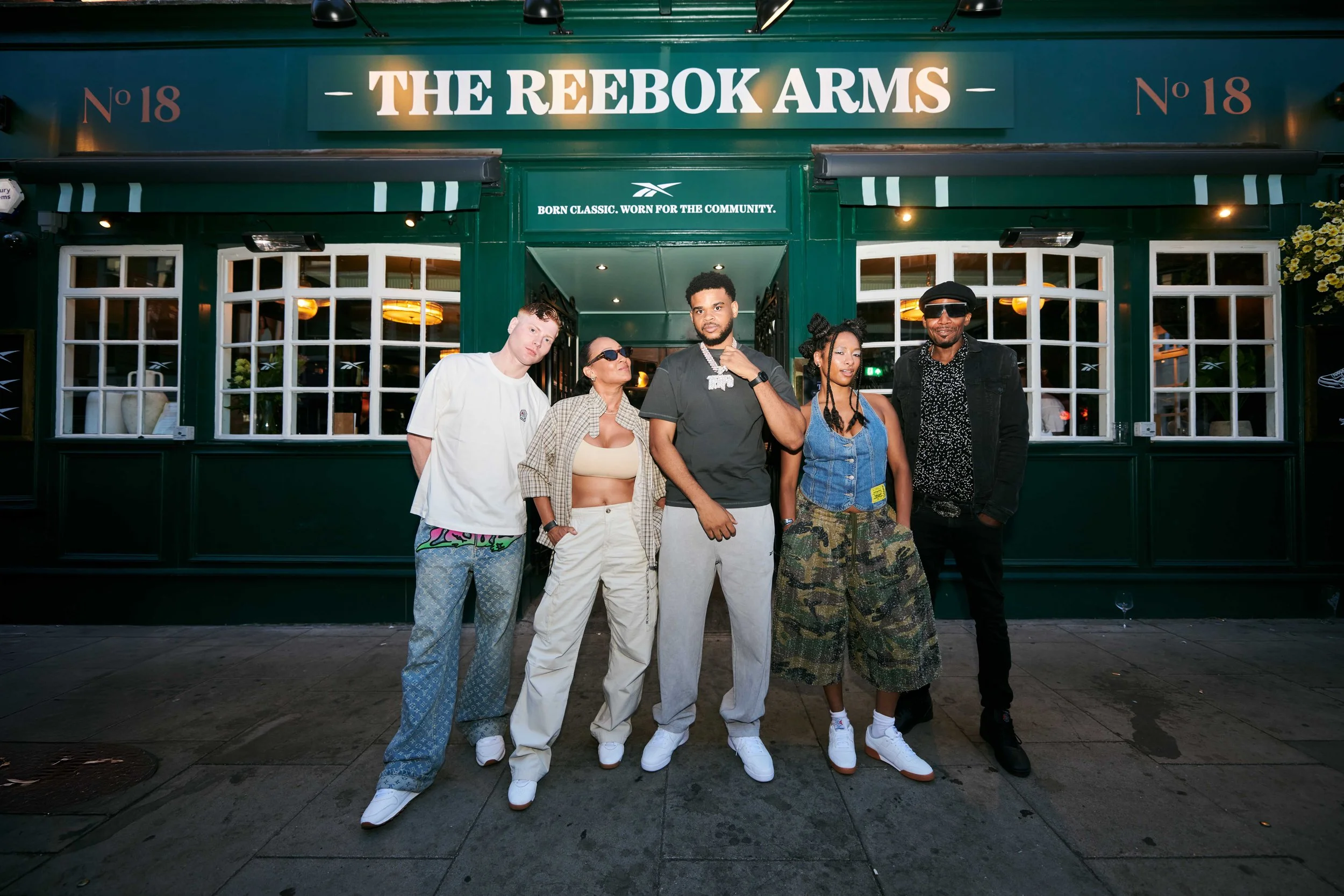 Five people standing in front of a green storefront with a sign that reads 'The Reebok Arms,' with the number 18 on either side of the sign. The group includes one person with light skin and four with darker skin, all dressed in casual, trendy clothing, and posing confidently for the photo.
