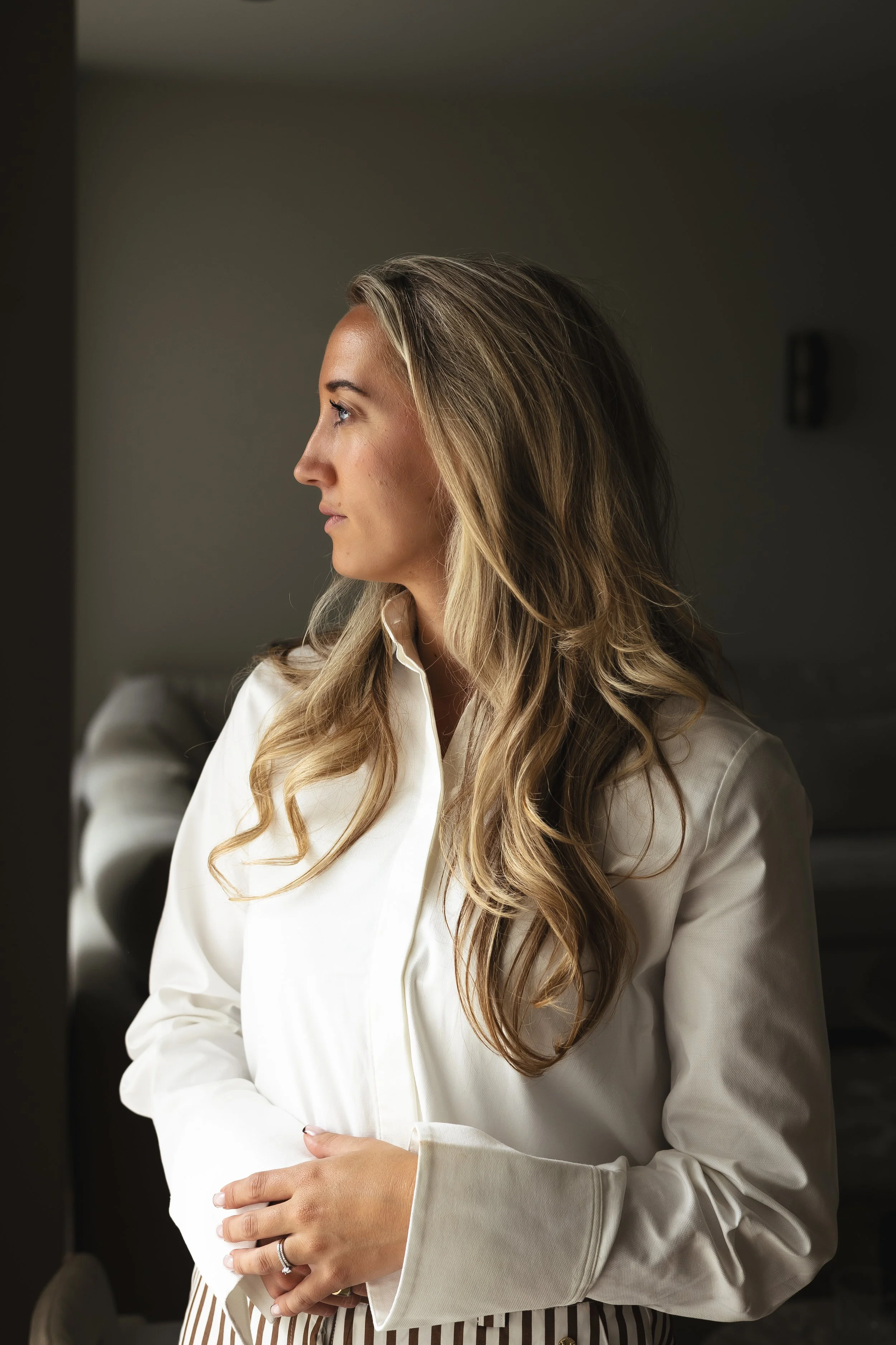 Een jonge vrouw met lang blond haar zit op een houndstooth patronen fauteuil in een modern interieur met witte muren en grote ramen. Ze draagt een bruin vest en witte broek.