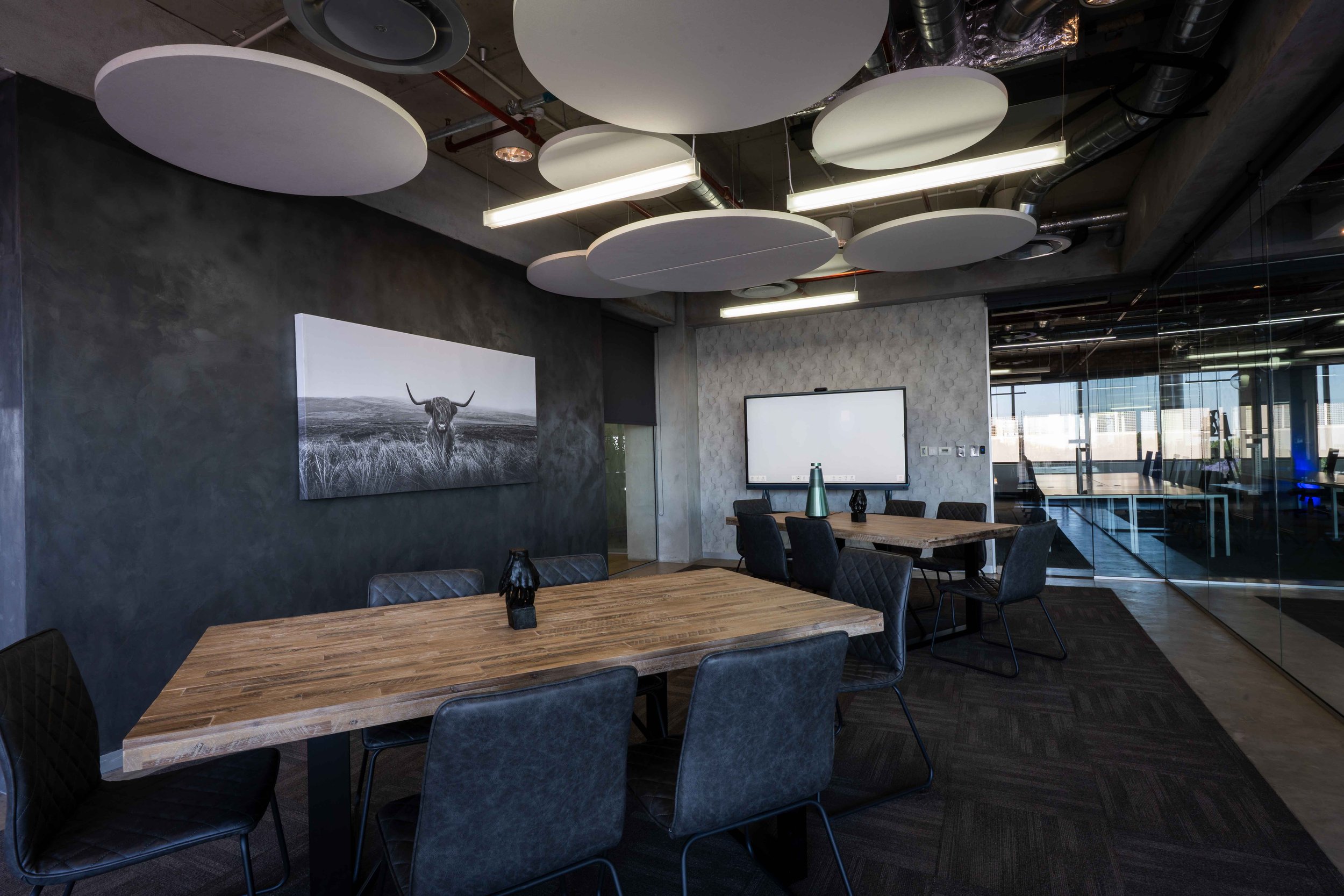 Modern office conference room with a wooden table, black chairs, a large whiteboard, and abstract ceiling panels in black and white, with floor-to-ceiling windows and cityscape view