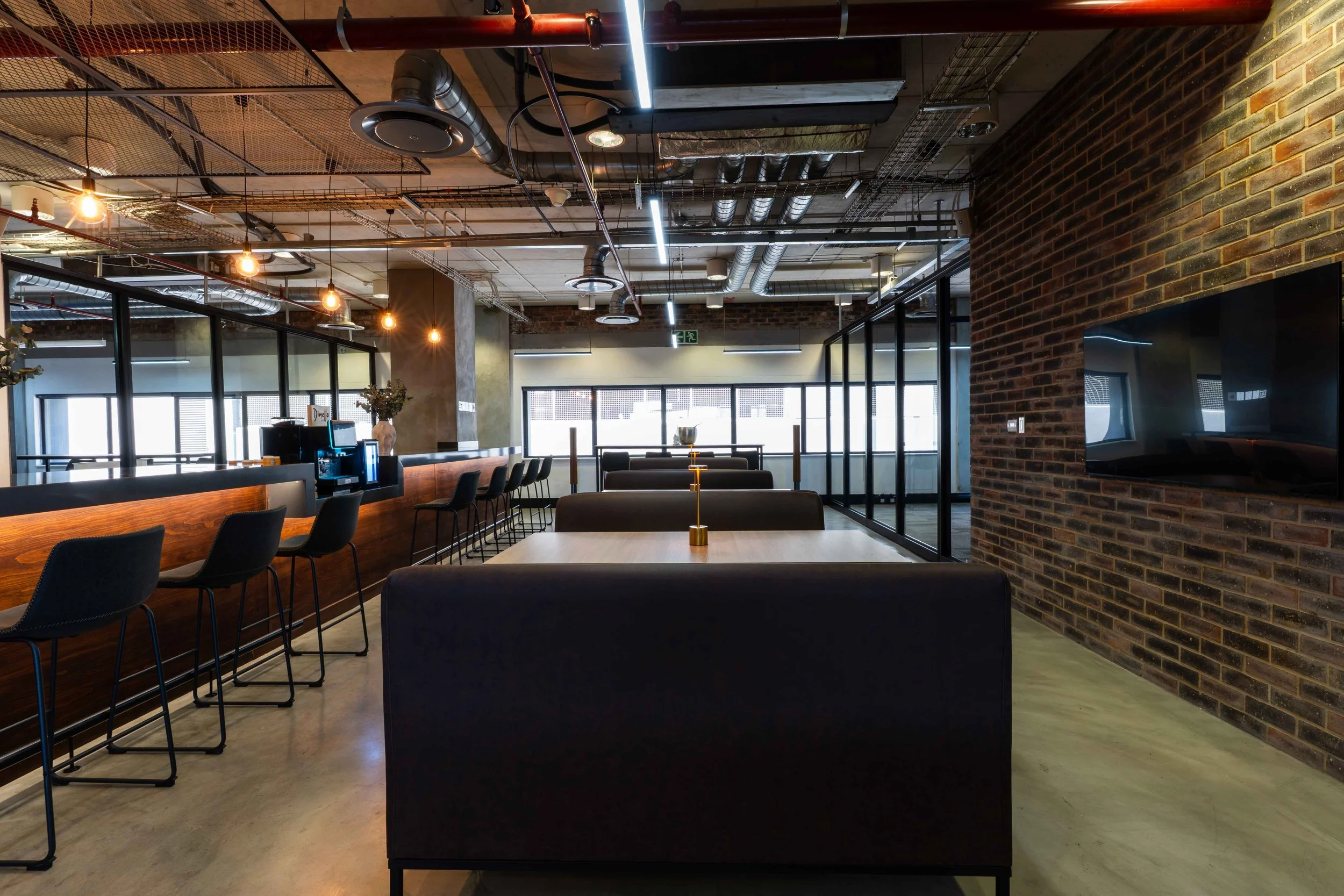 Modern cafe or coworking space with wooden and leather furniture, exposed ceiling ducts, large windows, and a brick wall with a mounted TV.