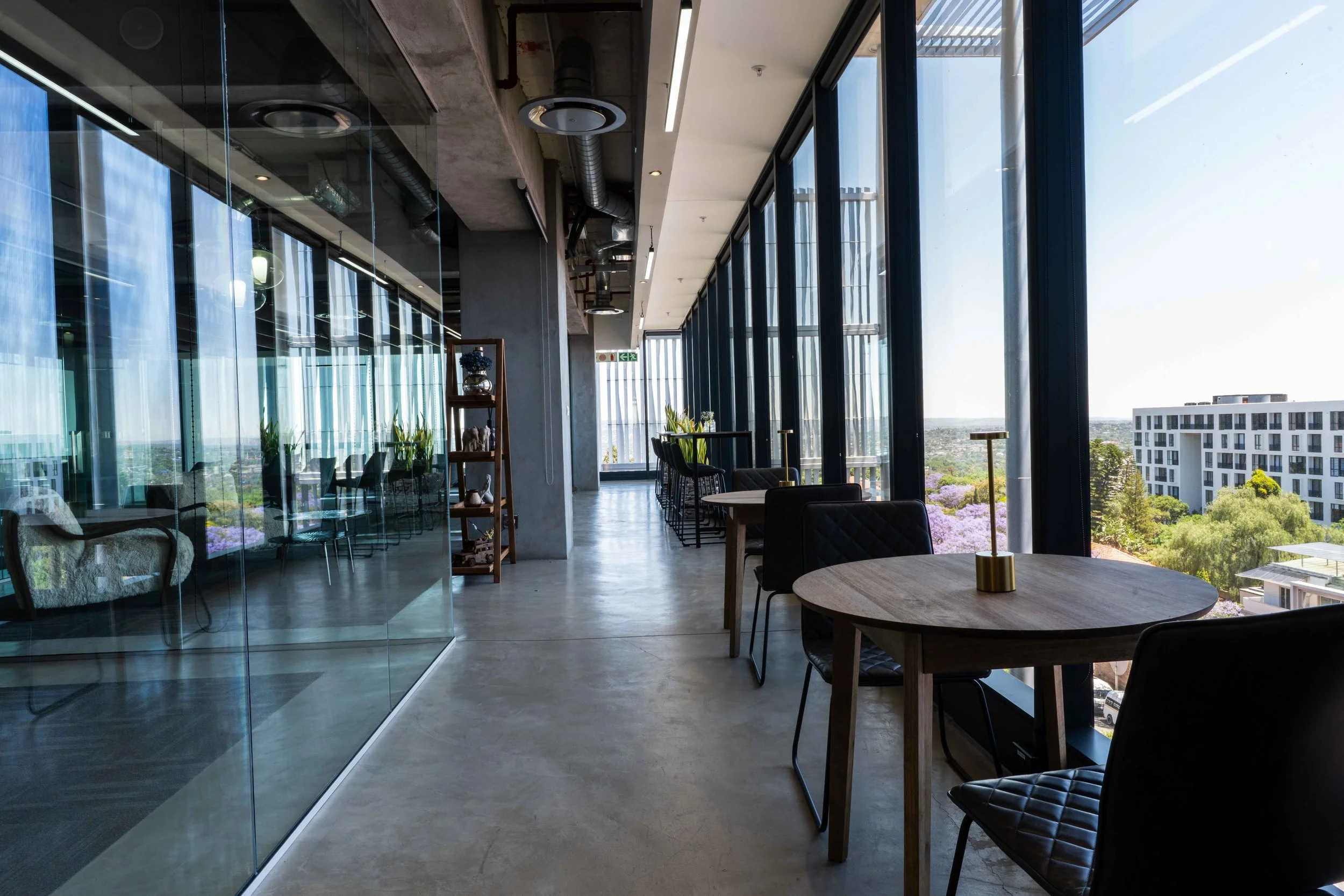 Interior of a modern high-rise cafe with floor-to-ceiling windows, black tiled chairs, wooden tables, and city view overlooking buildings and trees.