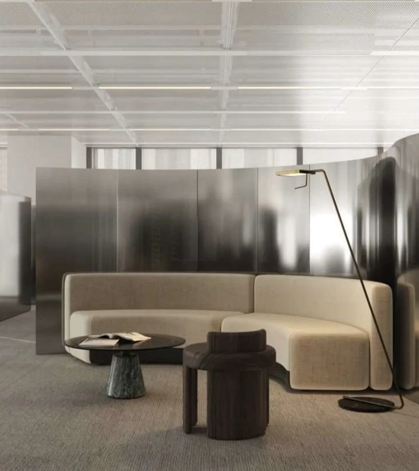 Modern airport lounge with curved beige sofa, black side table with open book, black ottoman, and tall floor lamp; metallic wall panels in the background and a high ceiling with linear lighting.